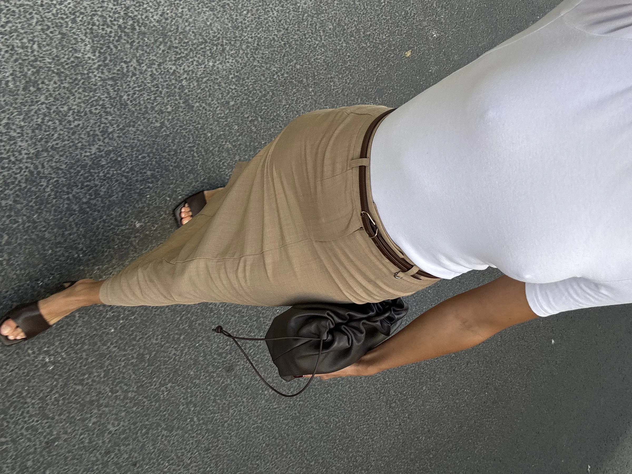 Overhead phone shot of a woman in a white tee, tan maxi skirt, brown belt and pouch bag on asphalt.