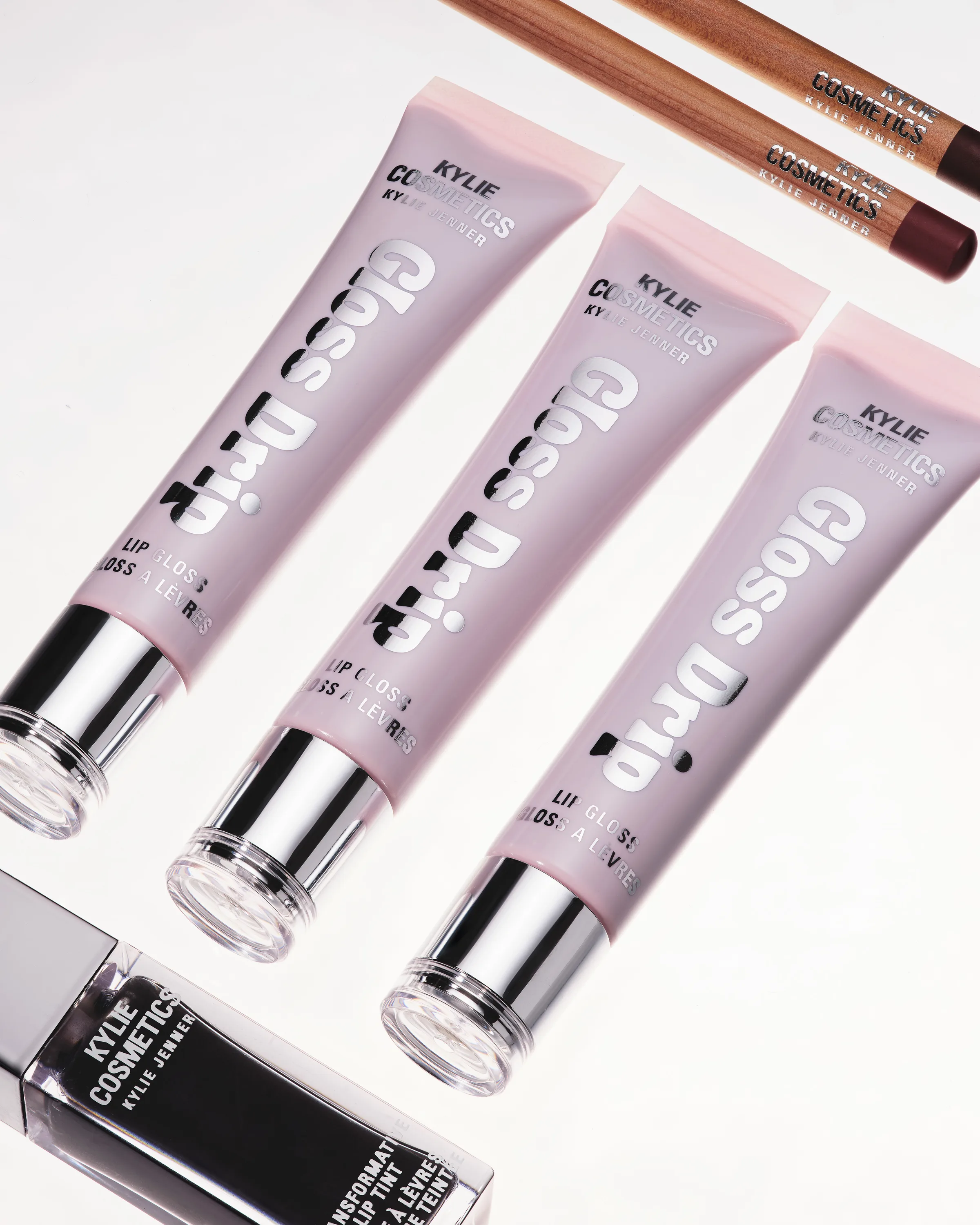 Three pink Gloss Drip lip gloss tubes with chrome caps, lip liners and a black lip tint on white, for Kylie Cosmetics.