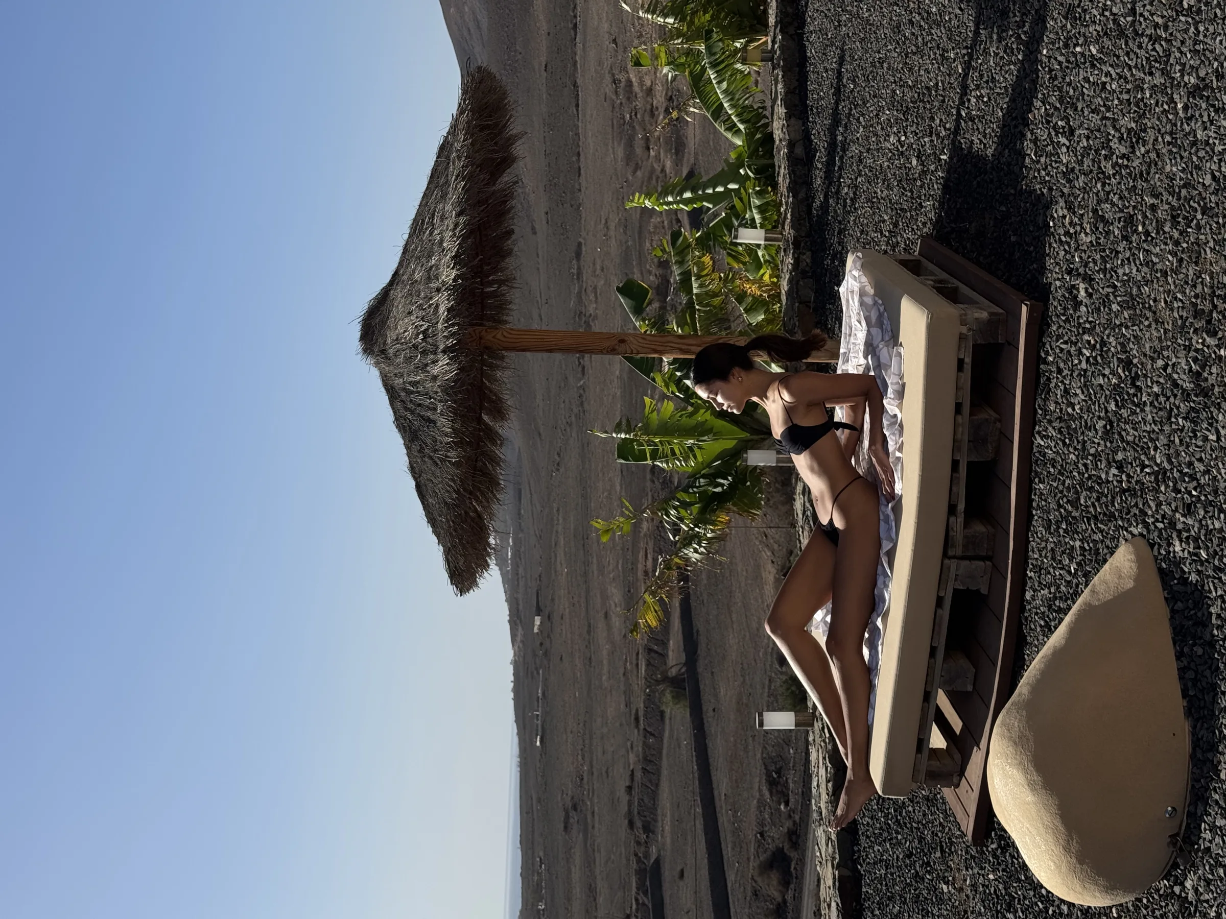 Woman in a black bikini sunbathing on a pallet daybed under a thatched umbrella in a volcanic desert landscape.