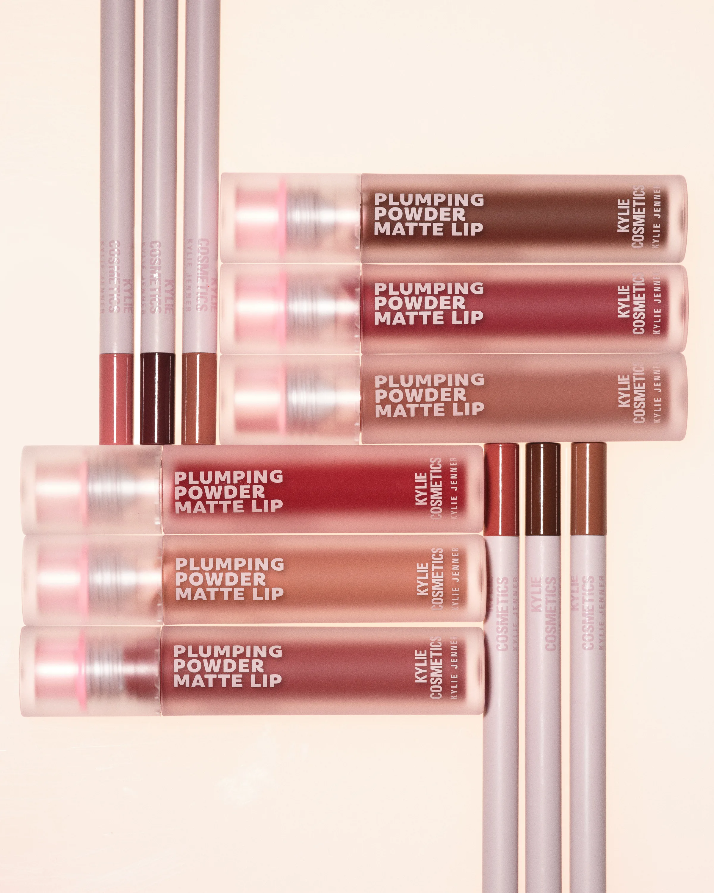 Plumping Powder Matte Lip tubes and lip liners in red and brown shades arranged on a peach backdrop, for Kylie Cosmetics.