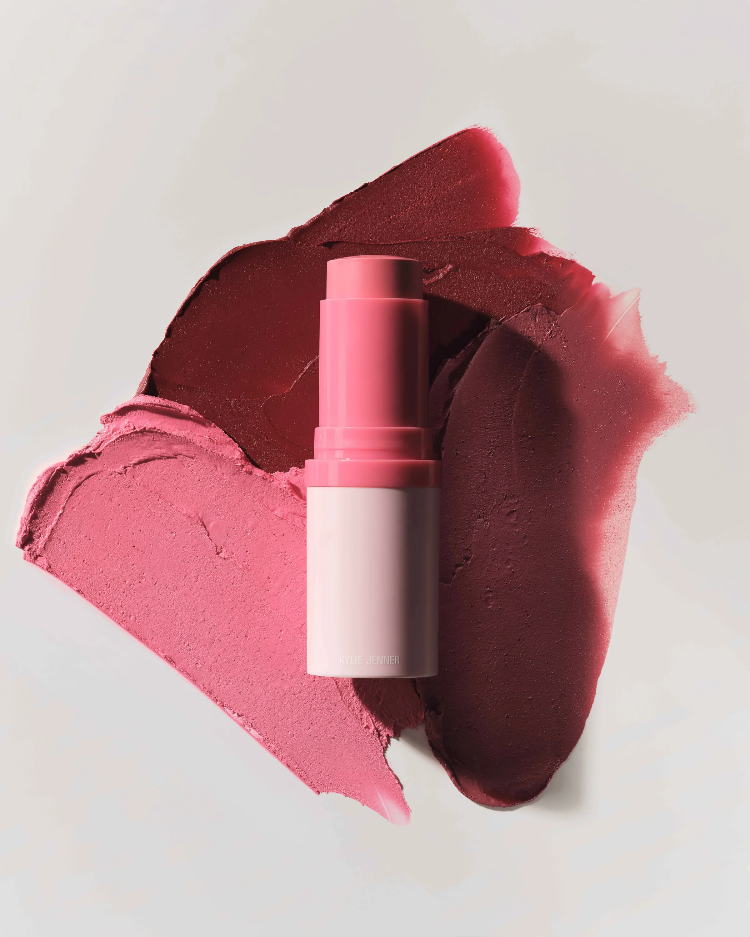 Pink blush stick lying on smeared pink and berry cream swatches against a pale grey background, for Kylie Cosmetics.