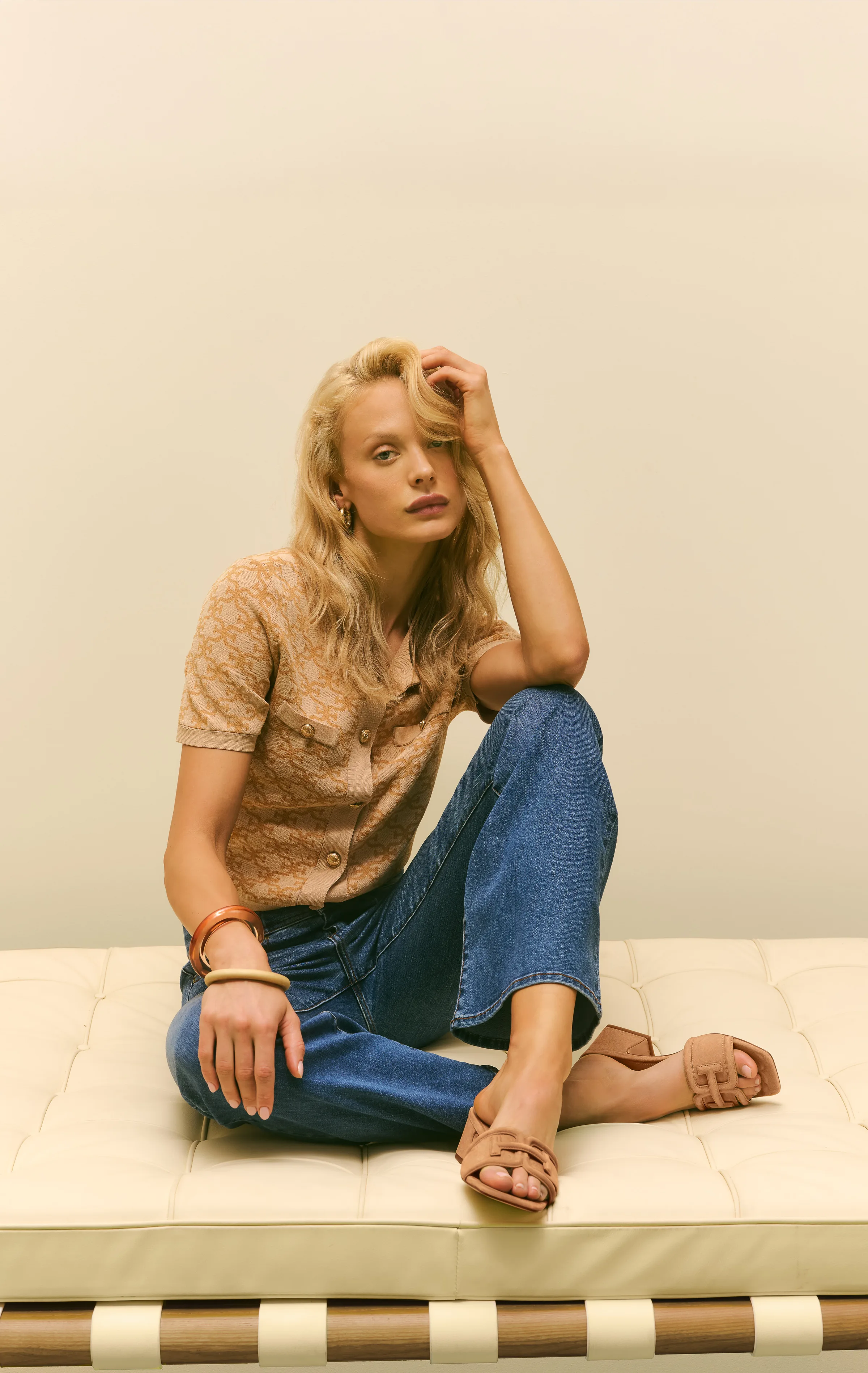 Blonde model in a tan knit top and jeans sits on a cream leather daybed wearing tan Waylon sandals, for Sam Edelman.