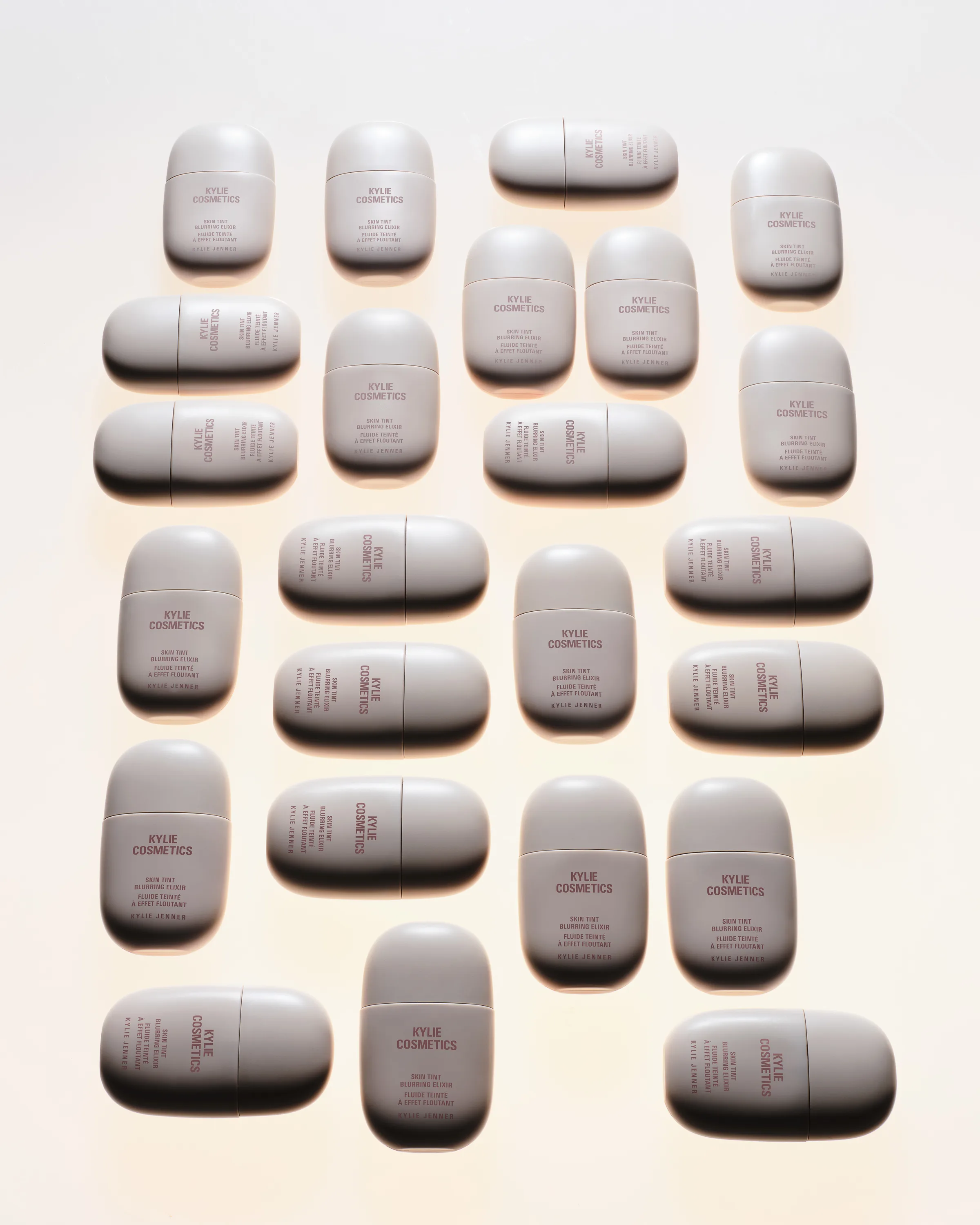 Rows of grey Skin Tint Blurring Elixir bottles arranged upright and sideways on a warm white background, for Kylie Cosmetics.