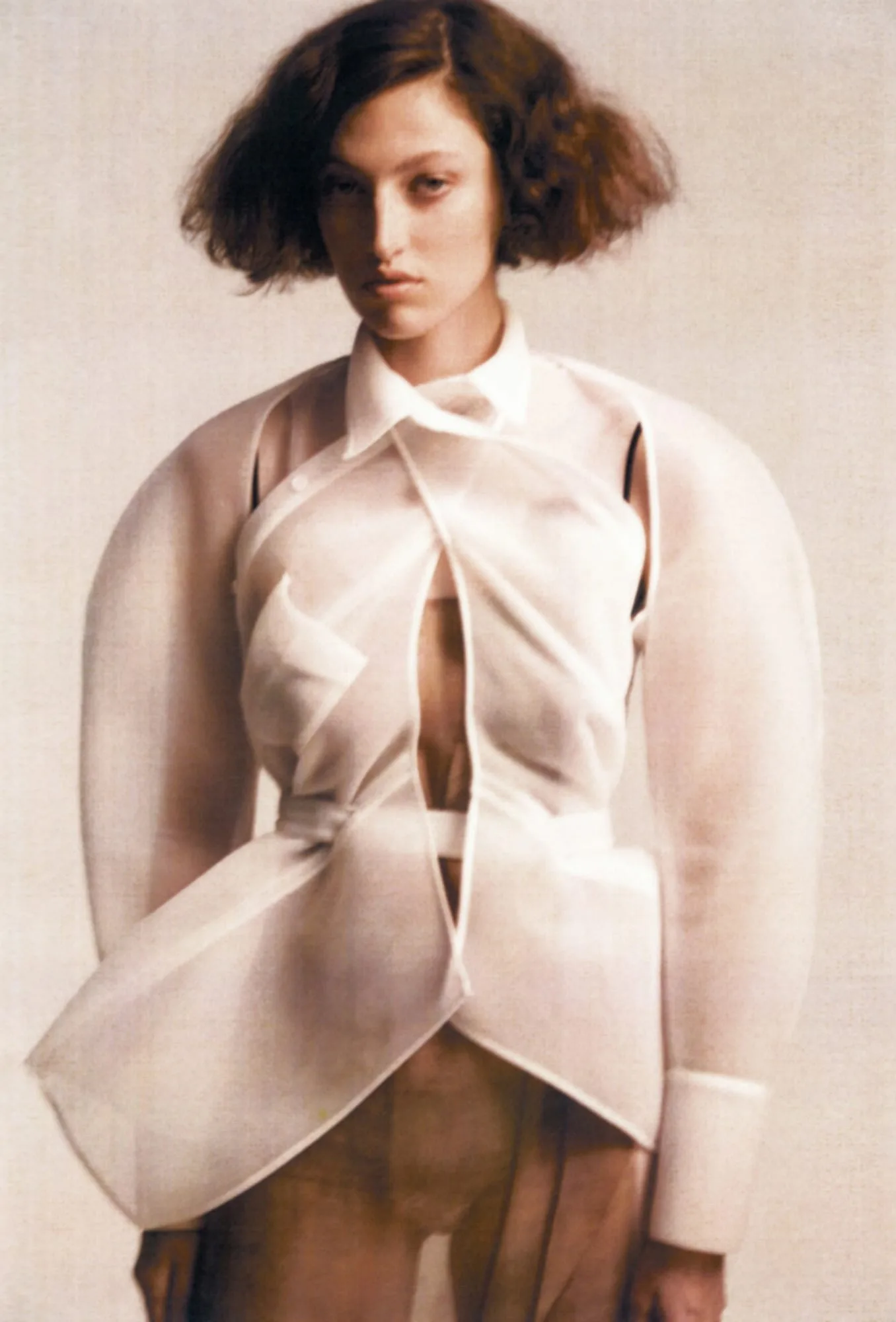 Model with bobbed curly hair in a sheer white sculptural jacket with rounded sleeves against a cream backdrop.