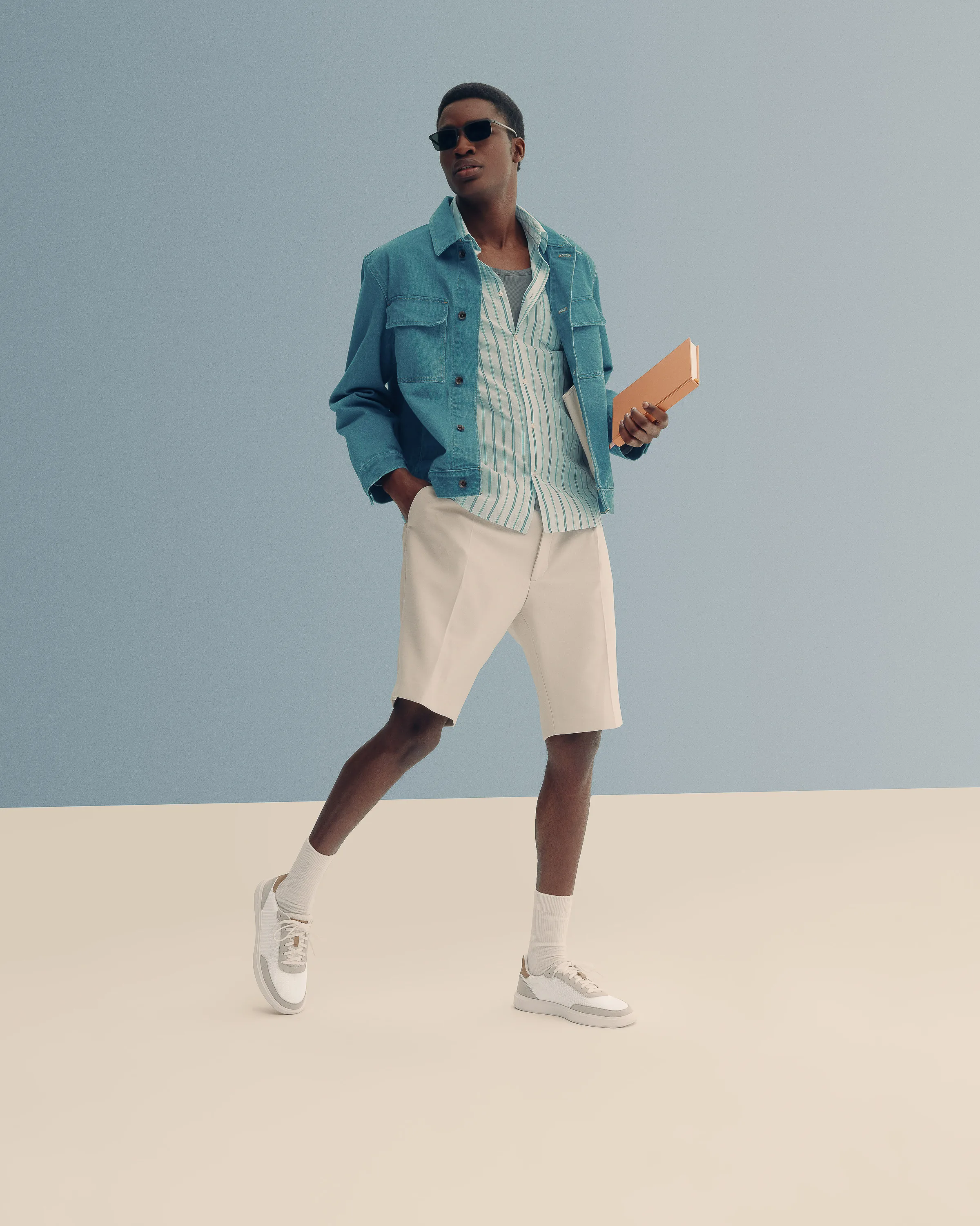 Man in teal denim jacket, striped shirt, cream shorts and white sneakers holding a tan folio on a blue set, for Cole Haan.