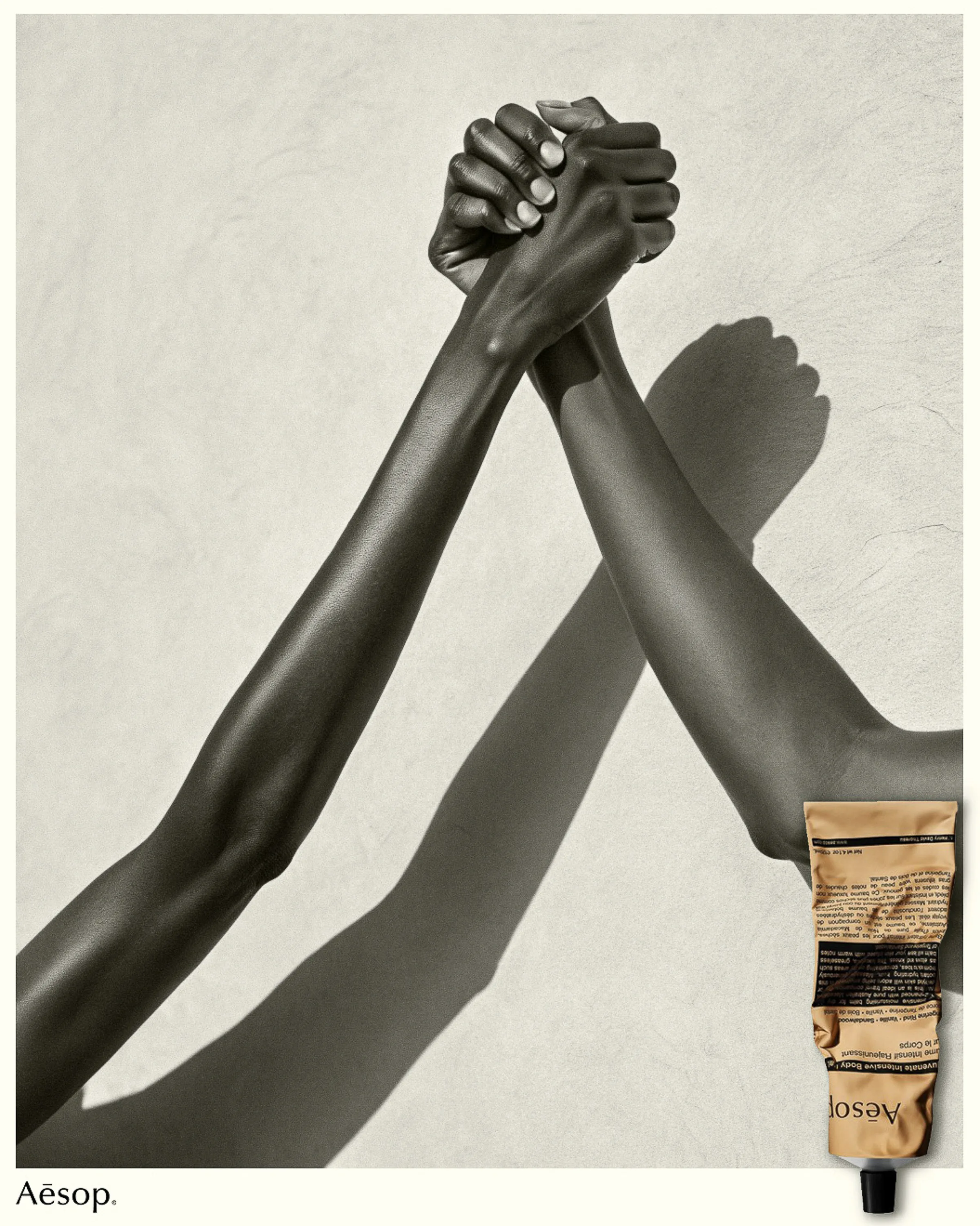 Two clasped arms raised against a sunlit plaster wall beside a tan tube of body balm. Aesop AI Concept.