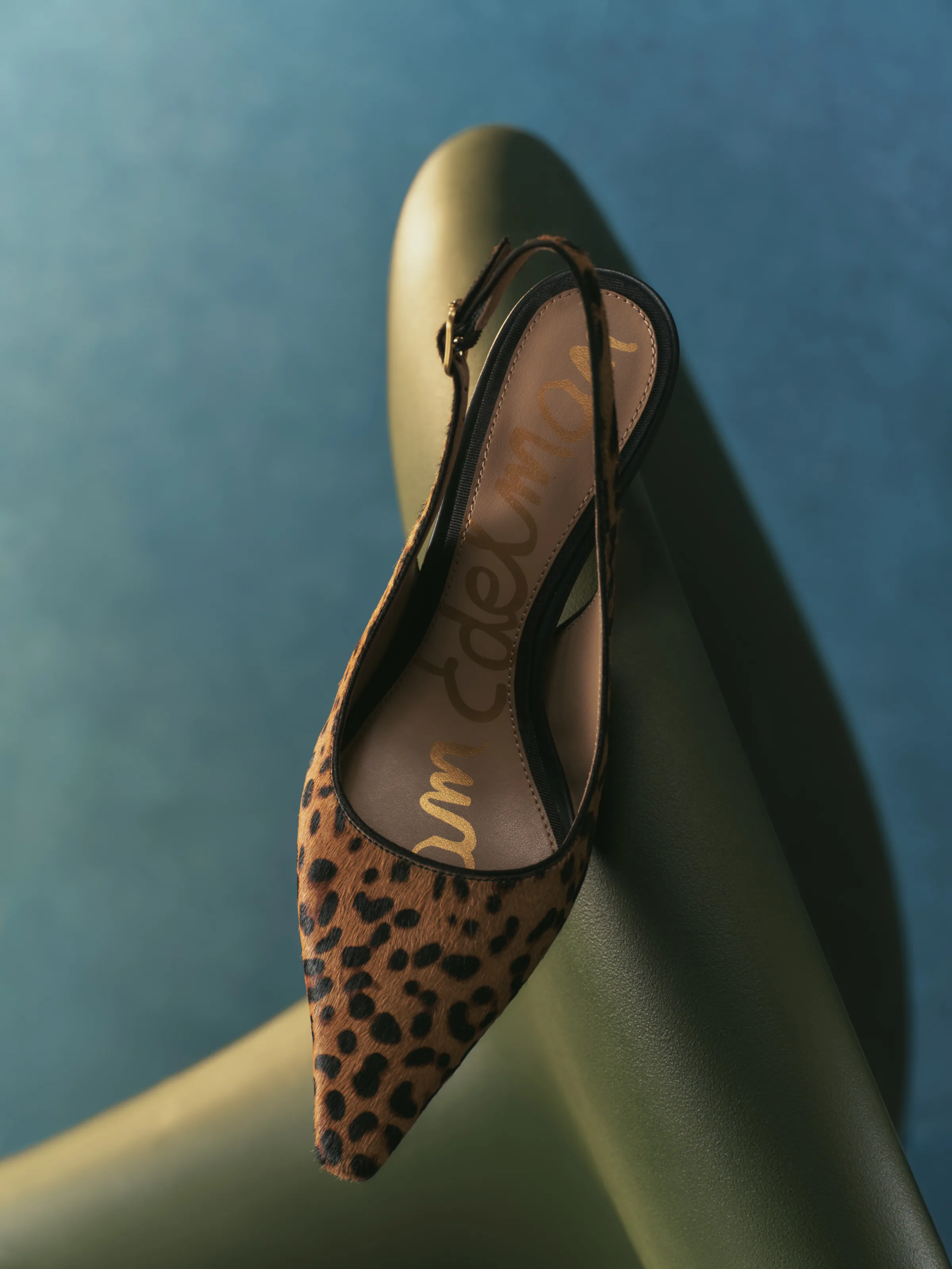 Leopard print calf hair slingback pump draped over a green leather chair against a teal wall, for Sam Edelman.