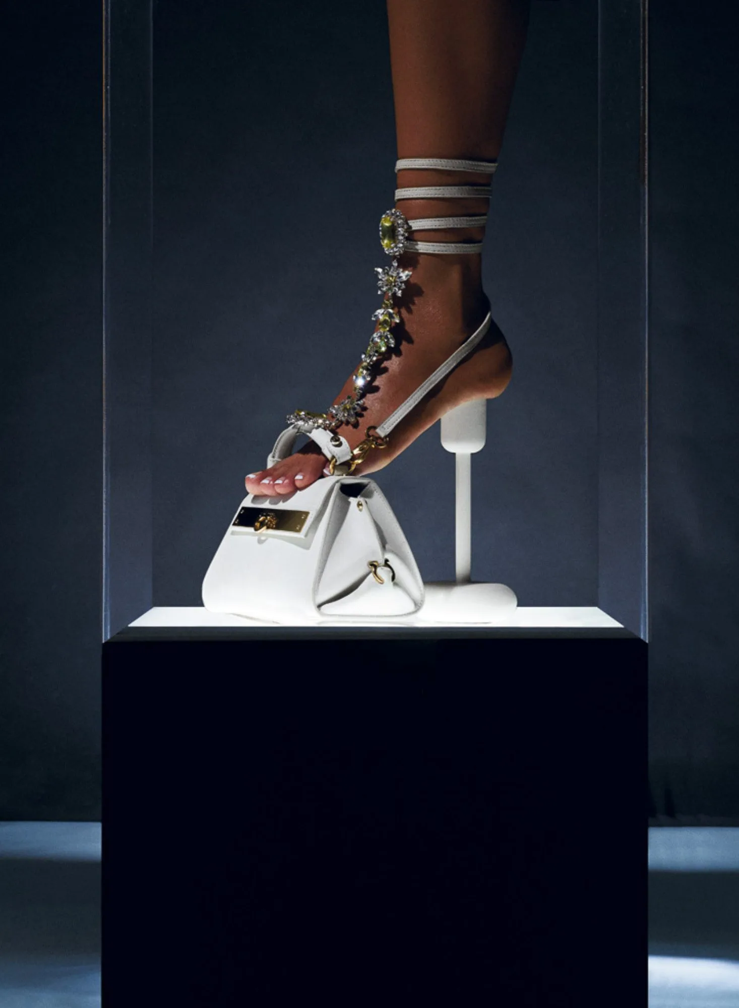Foot in a white jeweled strappy sandal resting on a white handbag atop a lit display plinth, for Interview Magazine.
