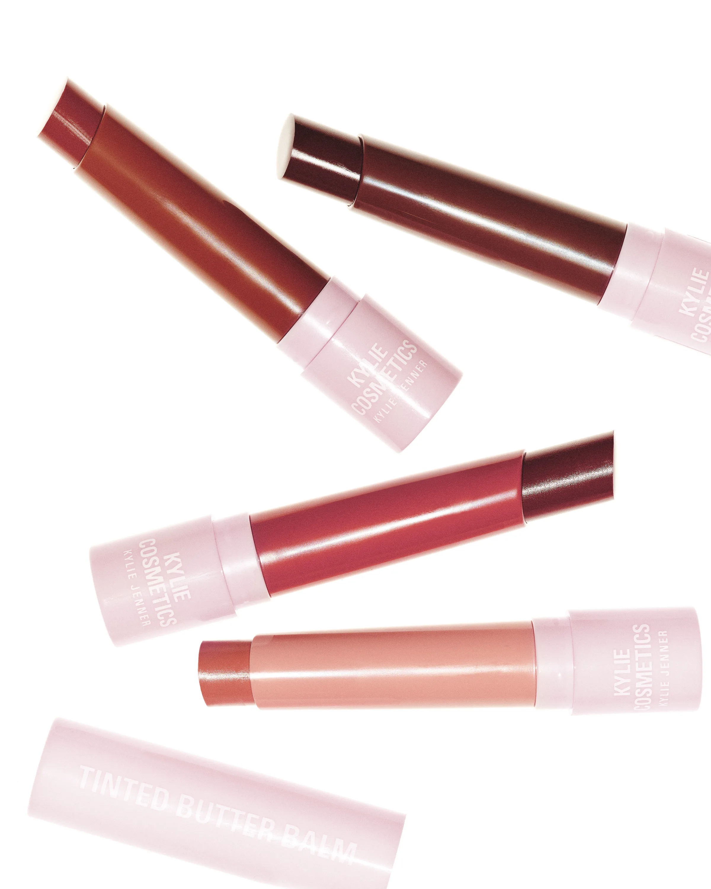 Four tinted butter balm sticks in red, berry and nude shades with pink caps scattered on white, for Kylie Cosmetics.