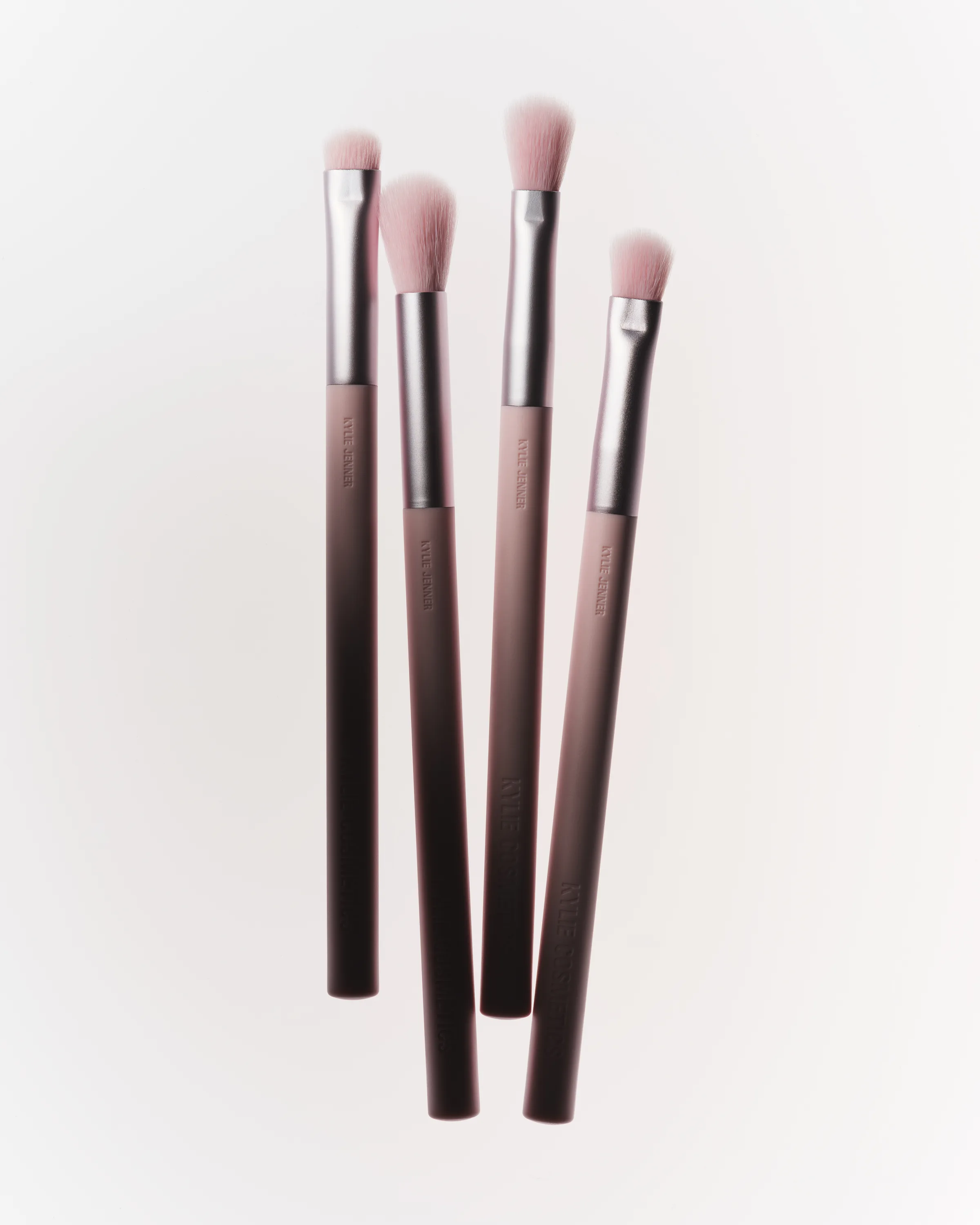 Four eyeshadow brushes with pink bristles and ombré mauve handles, arranged diagonally on white, for Kylie Cosmetics.