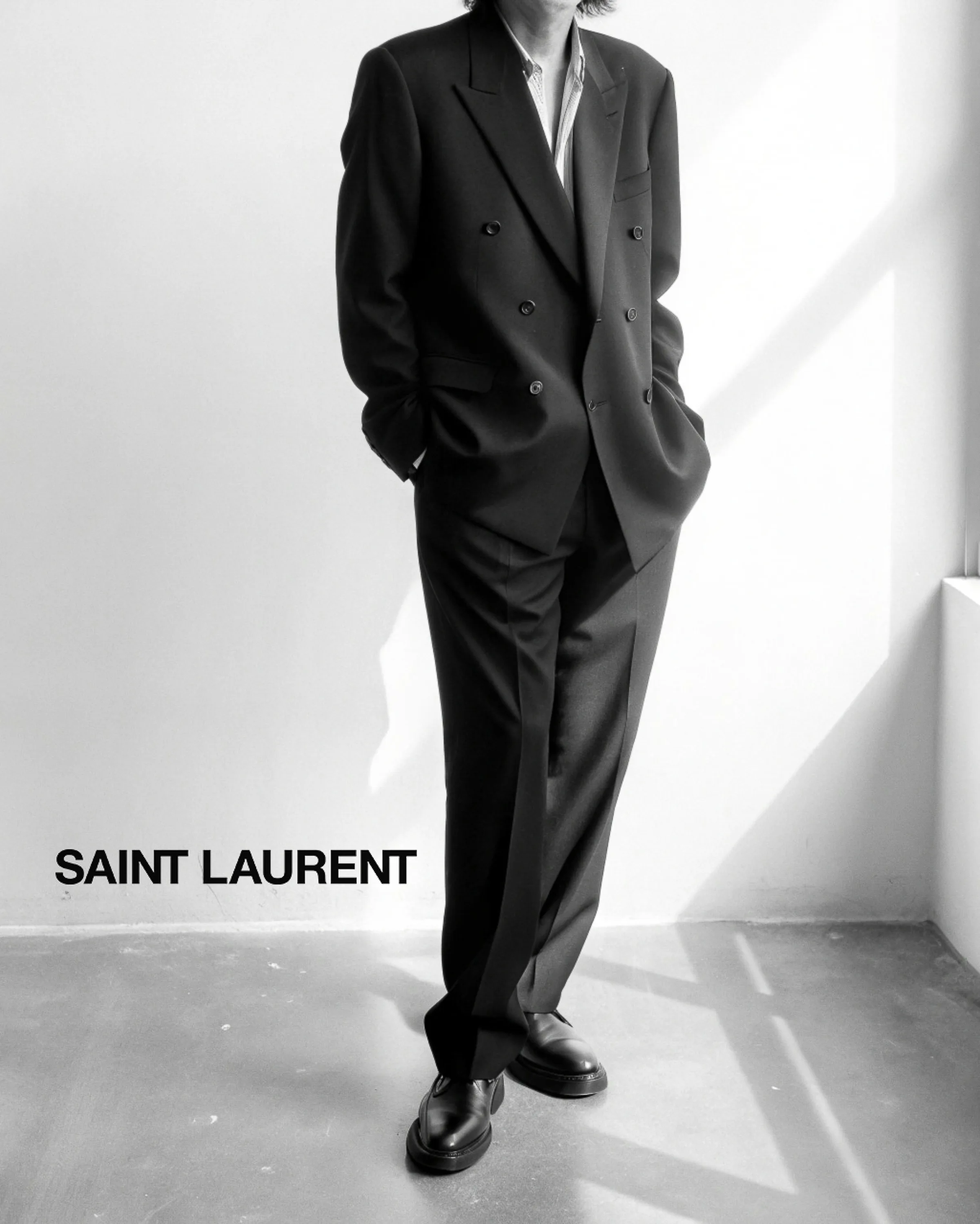 Black-and-white AI image of a model in a black double-breasted suit and loafers in a sunlit white room. Saint Laurent AI Concept.