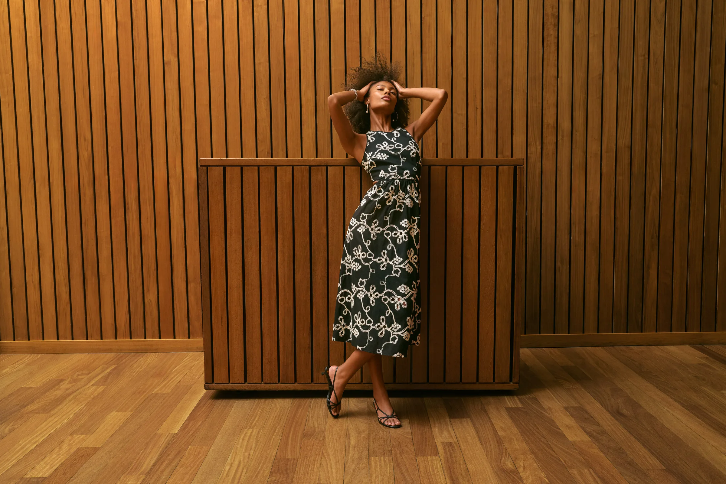 Model in a black dress with cream rope embroidery and strappy sandals poses against slatted wood panels, for Sam Edelman.