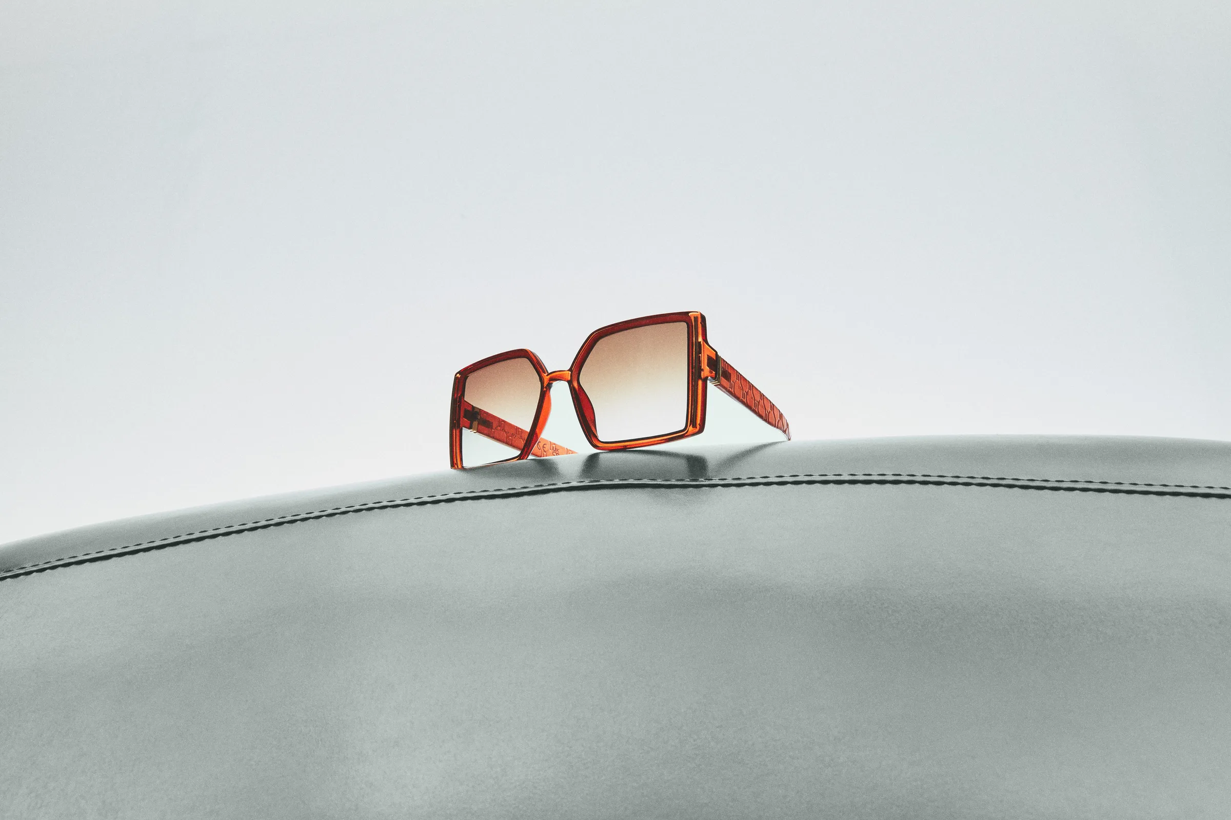 Oversized amber square sunglasses with gradient lenses resting on a grey stitched leather surface, for Sam Edelman.