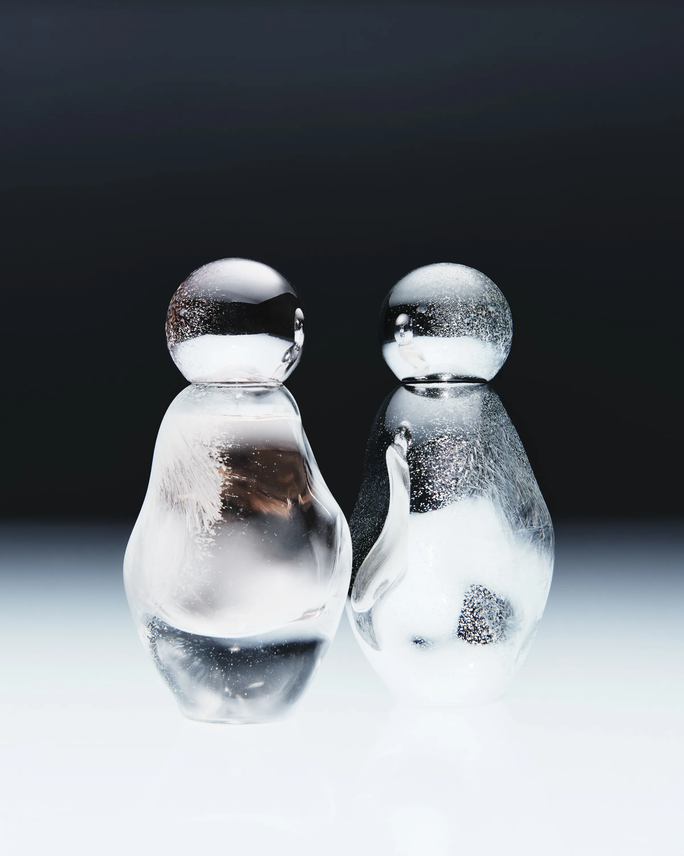 Two clear glass penguin figurines with silver glitter on a white surface against a dark backdrop, for Kylie Cosmetics.