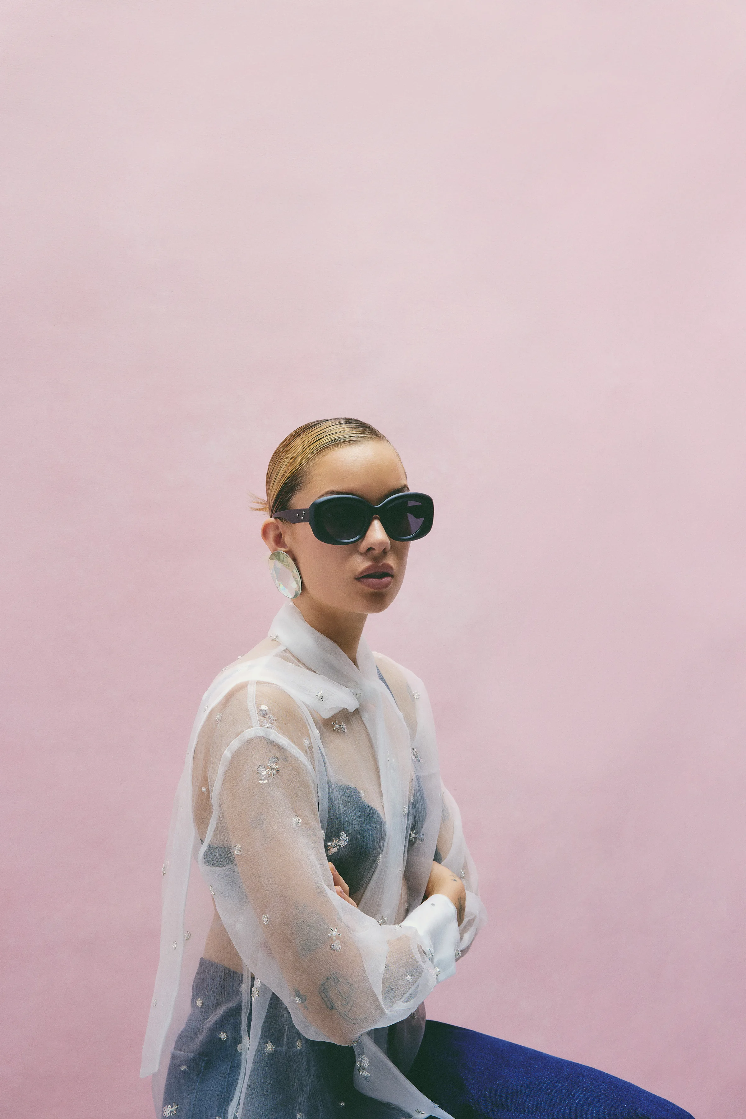 Model in a sheer embellished organza blouse, black sunglasses and statement earring seated against a pink wall, for Sam Edelman.