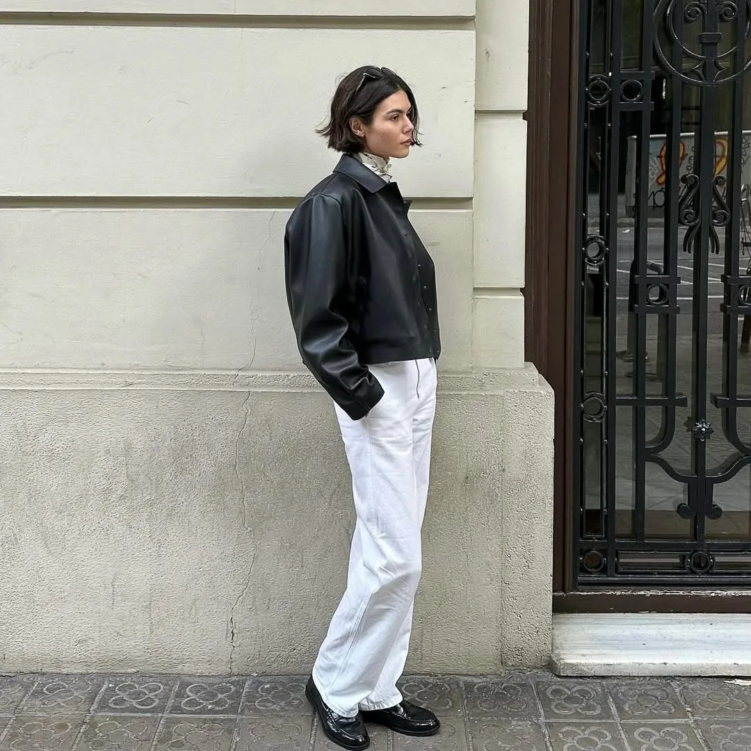 Woman in a cropped black leather jacket, white trousers and loafers leaning against a cream building beside an iron door.