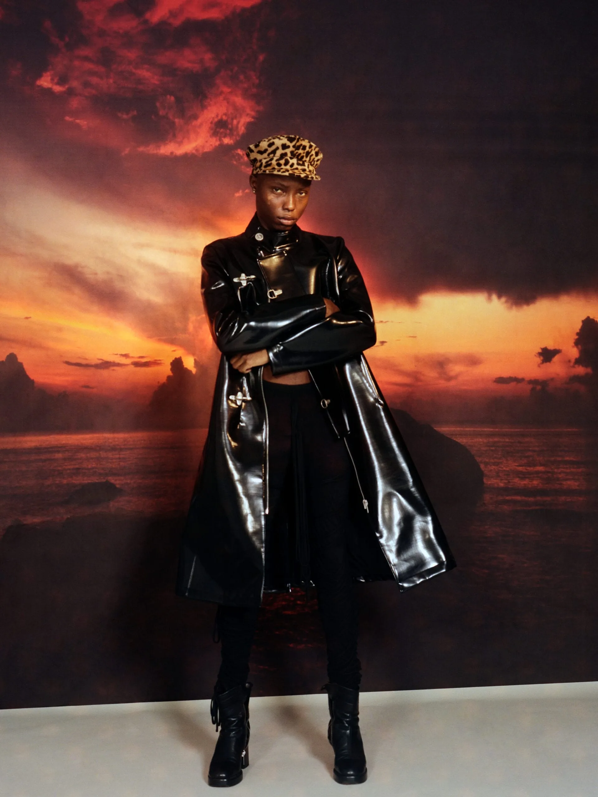 Model in a black patent trench coat, leopard cap and boots, arms crossed before a sunset seascape backdrop, for Office Magazine.
