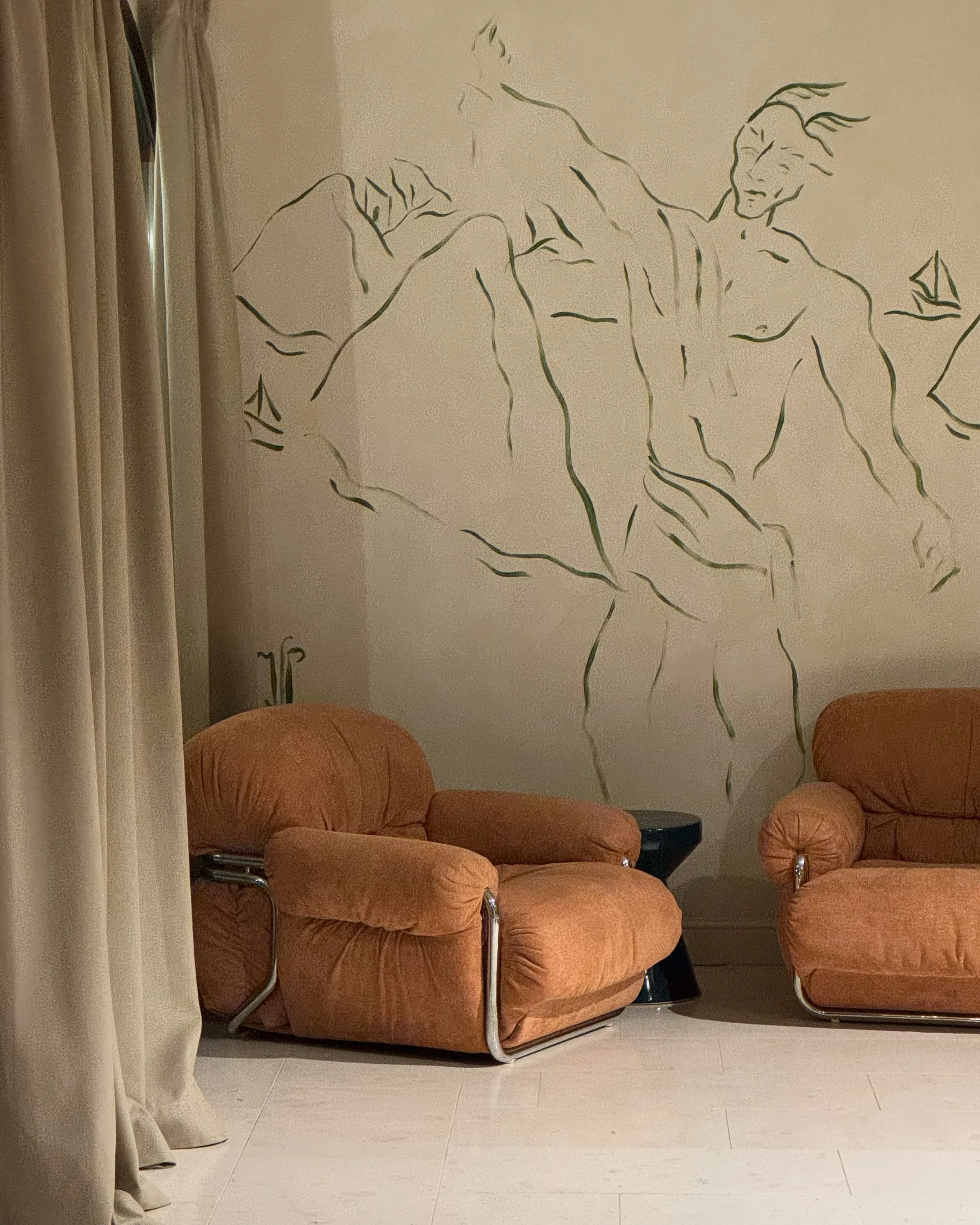 Two rust bouclé armchairs with chrome frames beside a black side table, set before a hand-drawn figure mural and linen curtains.