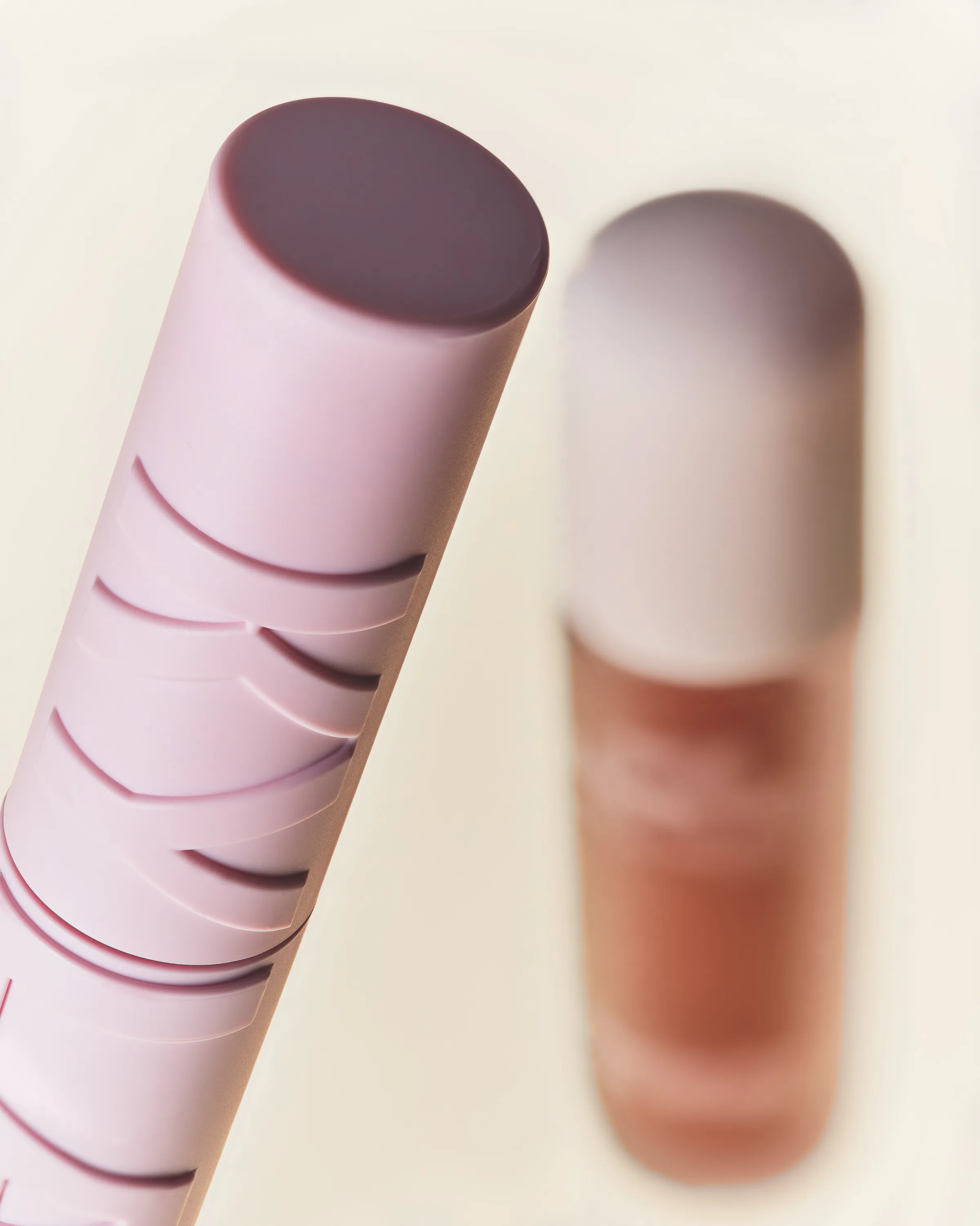 Close-up of a pink sculpted mascara tube with a blurred brown lip gloss behind on cream, for Kylie Cosmetics.