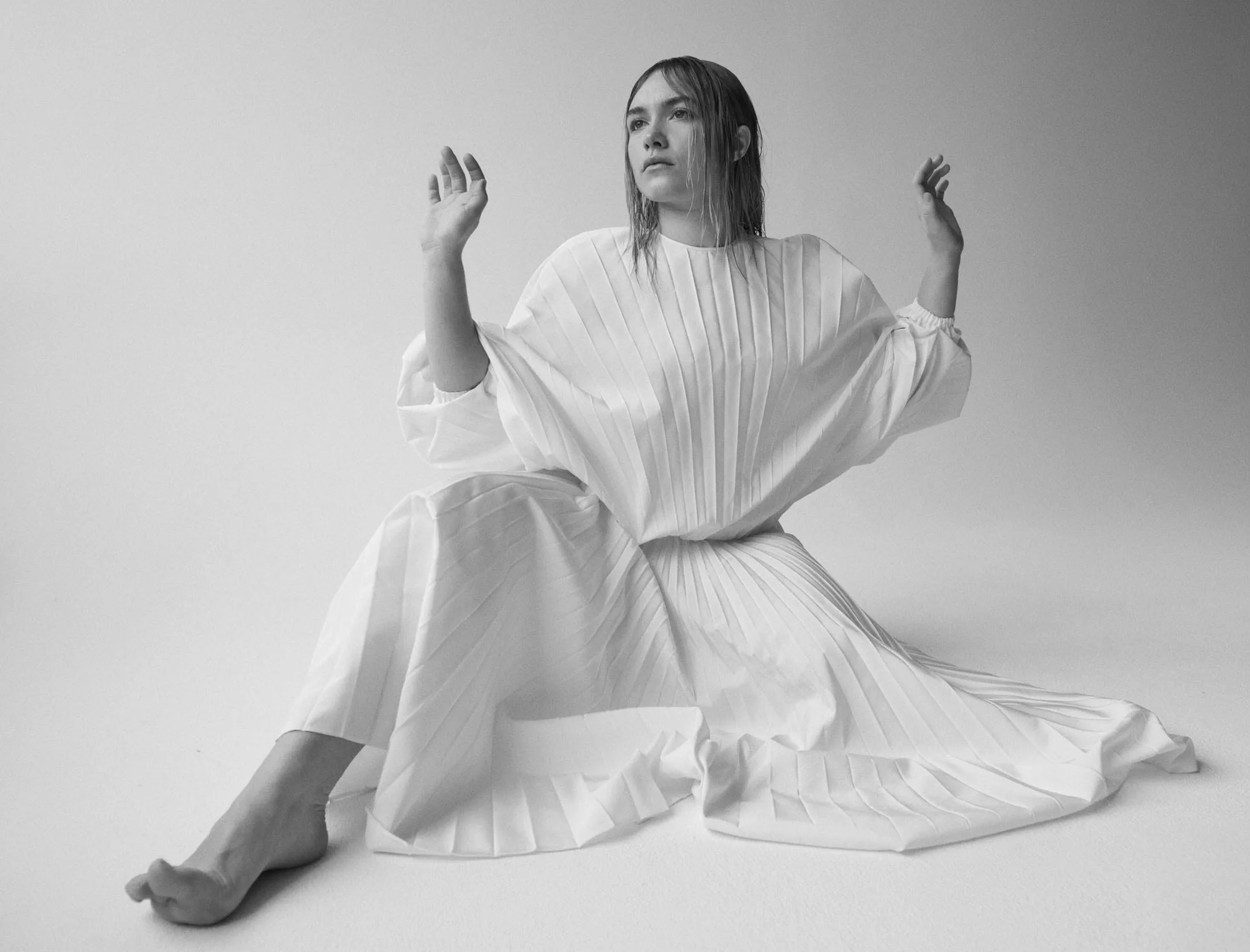 Black and white editorial of a barefoot model seated on the floor in a white pleated dress, hands raised, for The Last Magazine.