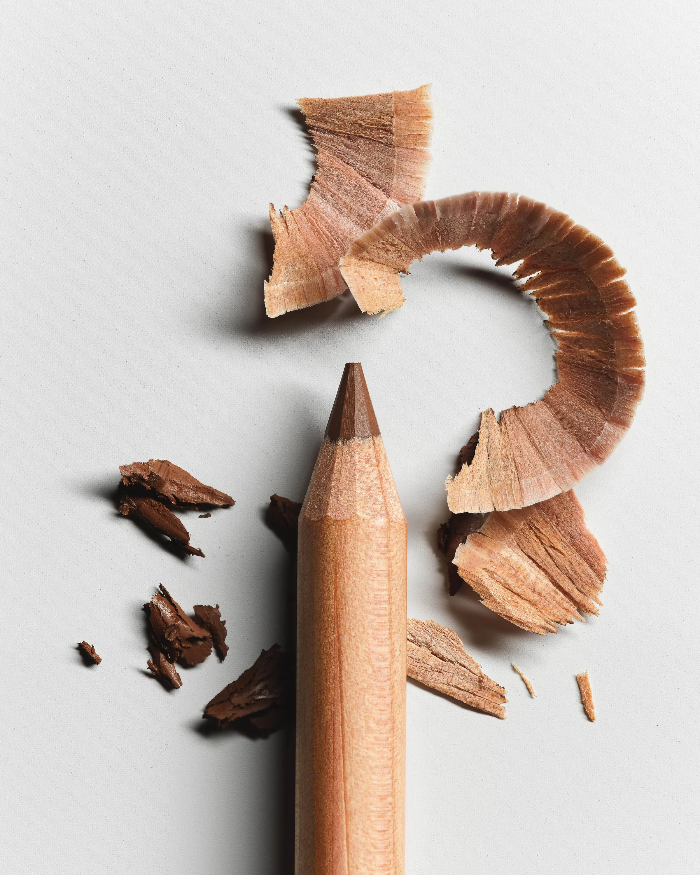 Still life of a sharpened brown lip liner pencil with curled wood shavings and broken pigment on white, for Kylie Cosmetics.