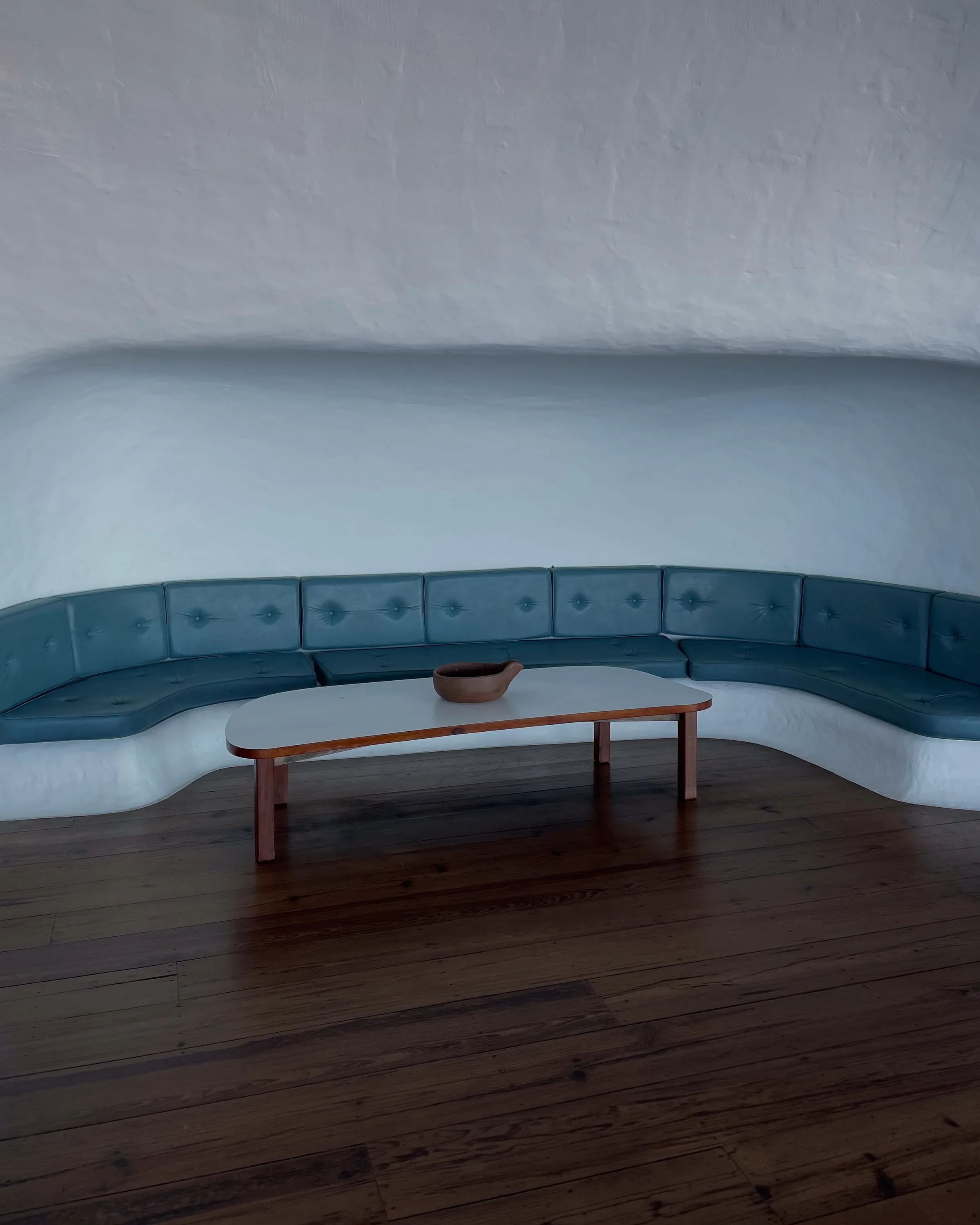 Curved teal leather banquette in a white plaster alcove with a wooden coffee table and clay bowl on dark plank floors.