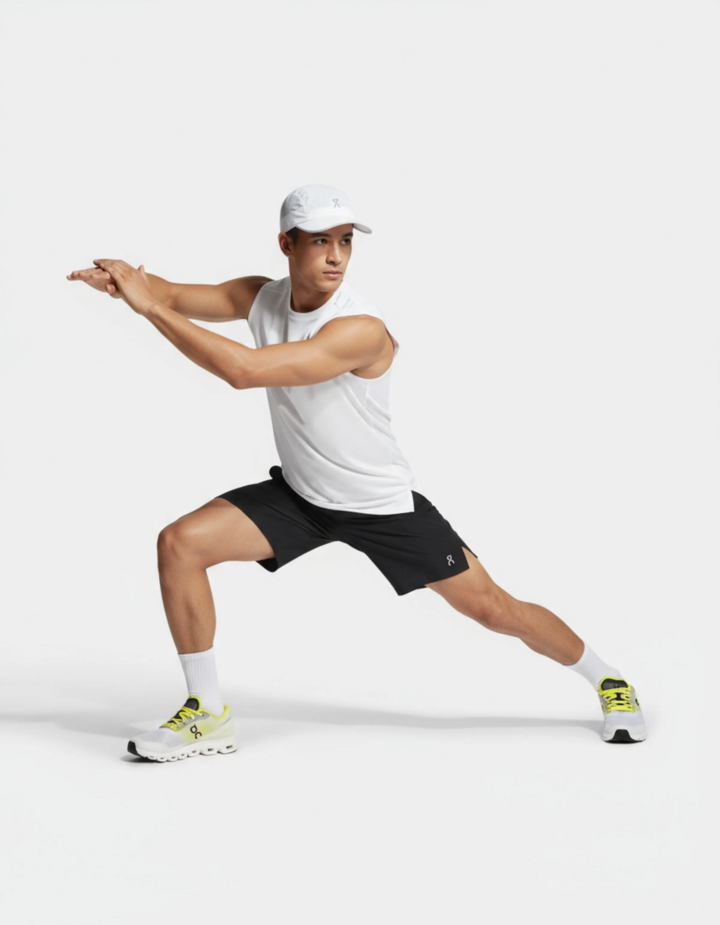 Male model in a side lunge wearing a white tank, cap, black shorts and lime-accented sneakers on a white backdrop. On AI Concept.