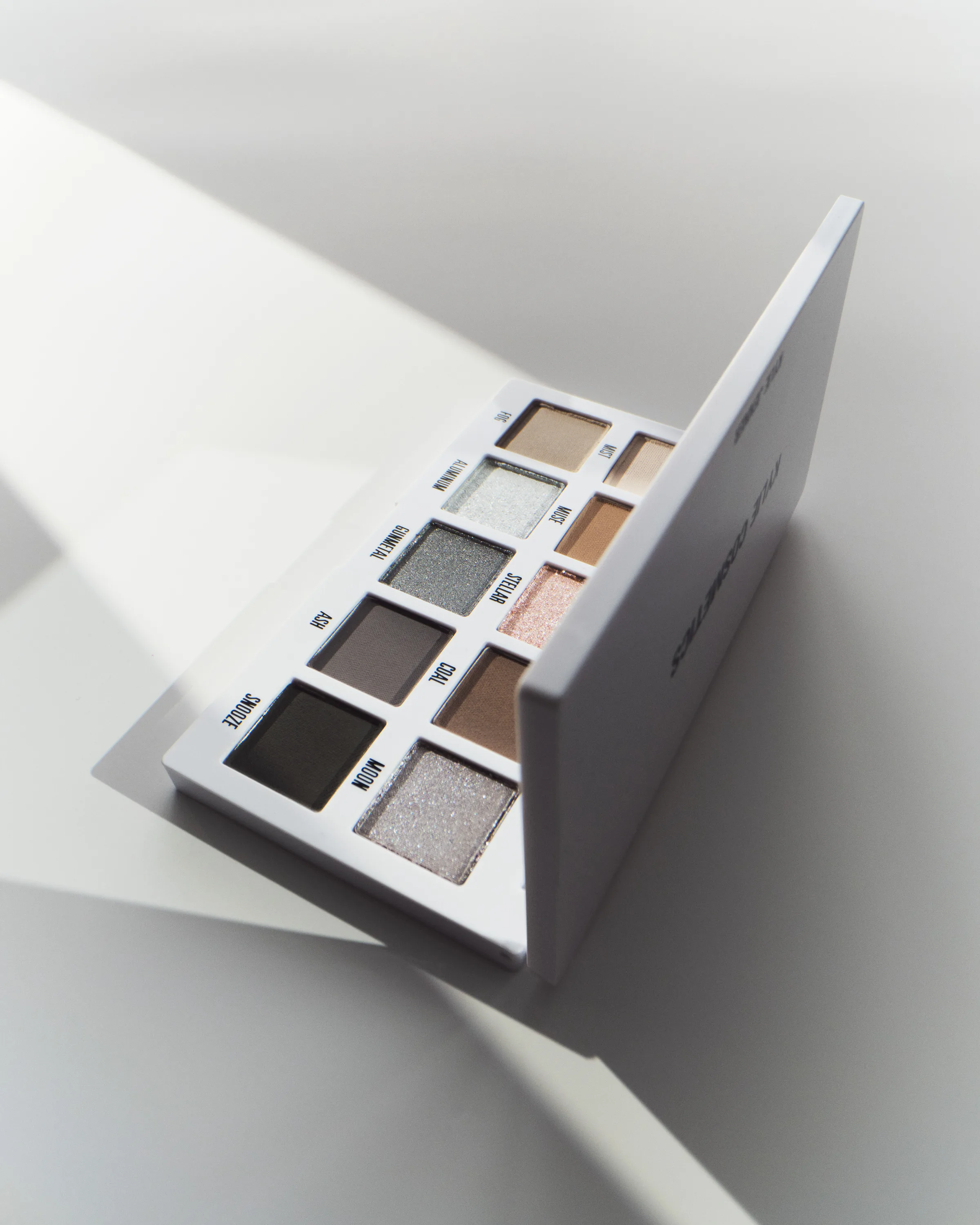 Open white eyeshadow palette of smoky grey, silver and taupe shades casting hard sunlight shadows, for Kylie Cosmetics.