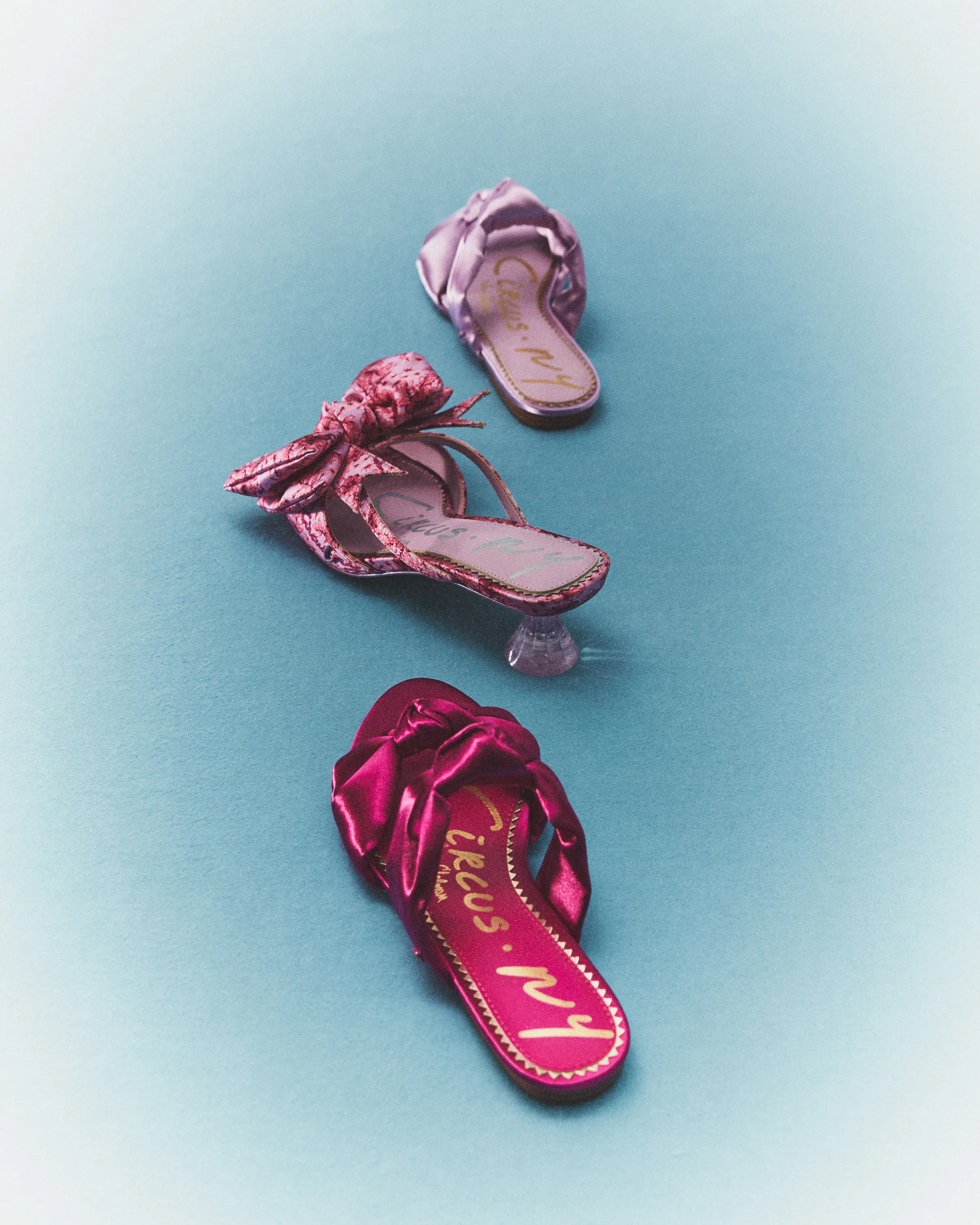 Pink and lilac satin bow slides and a clear-heeled mule on a teal backdrop, for Circus NY.