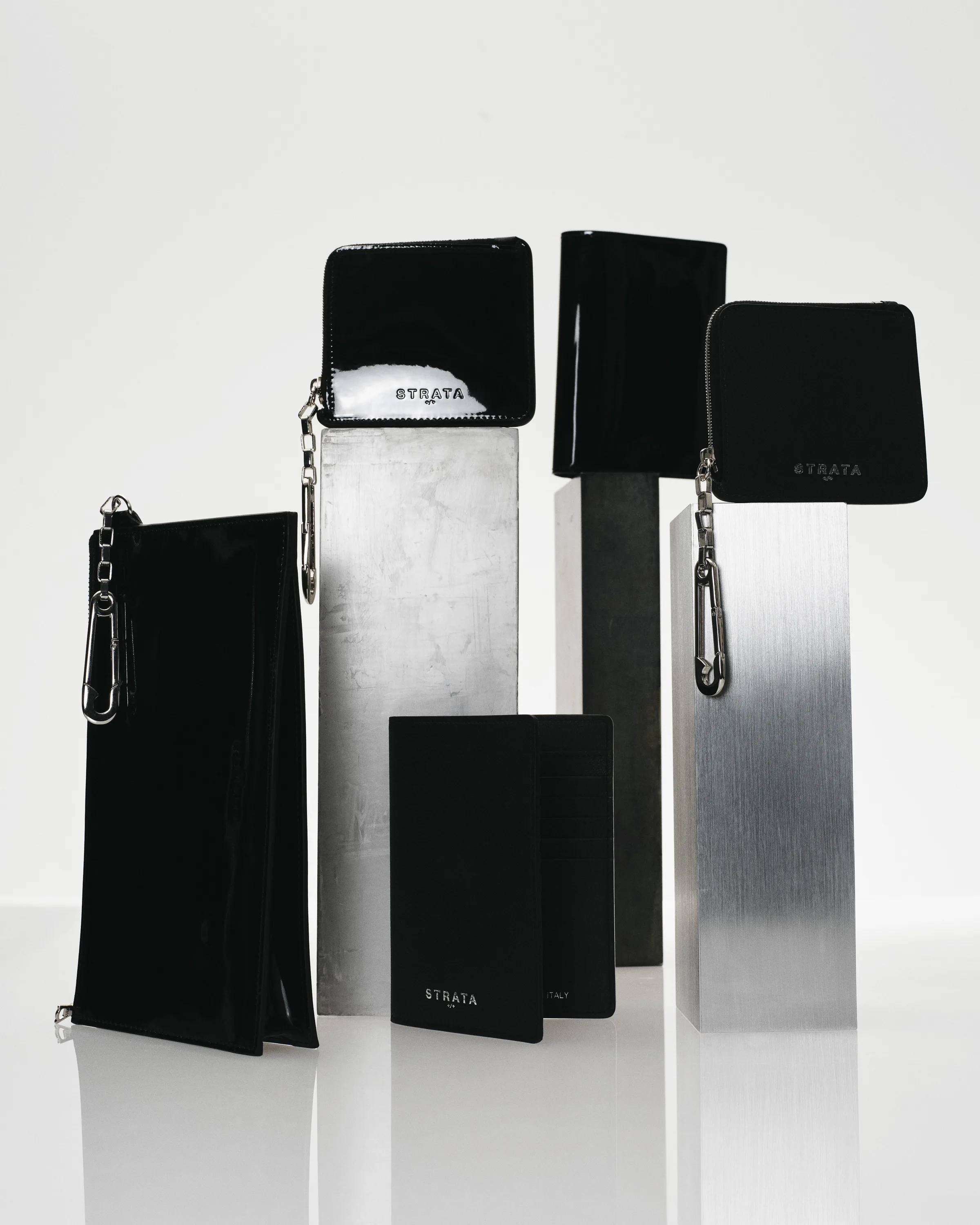 Black patent and matte leather wallets and pouches with silver clasps on brushed metal plinths, for Strata.