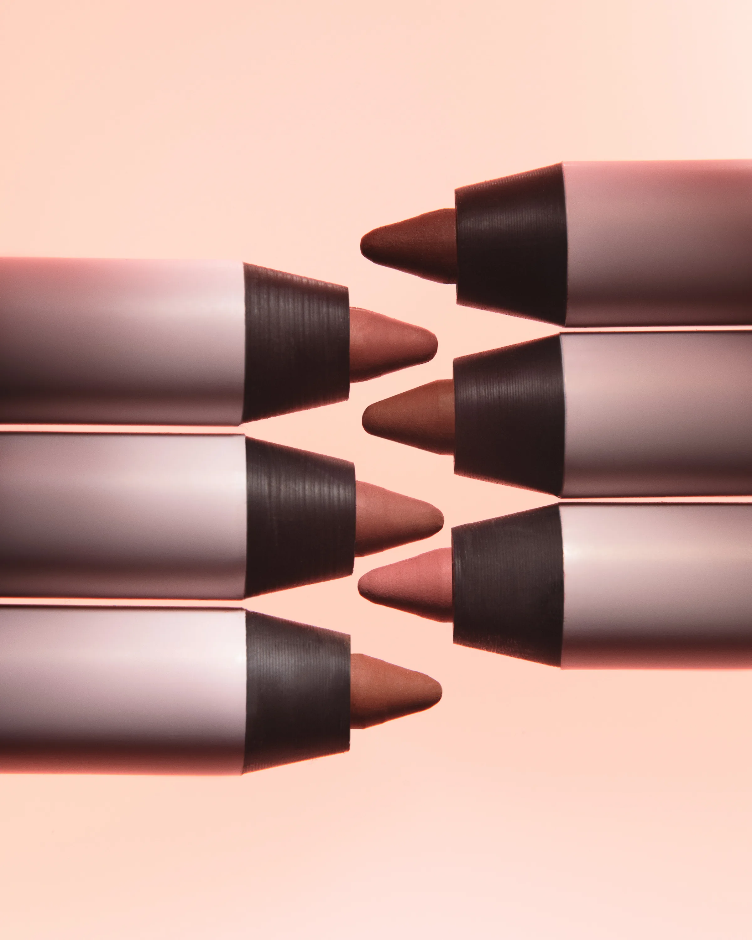 Six nude and brown lip liner pencils interlocking tip to tip on a soft peach background, for Kylie Cosmetics.