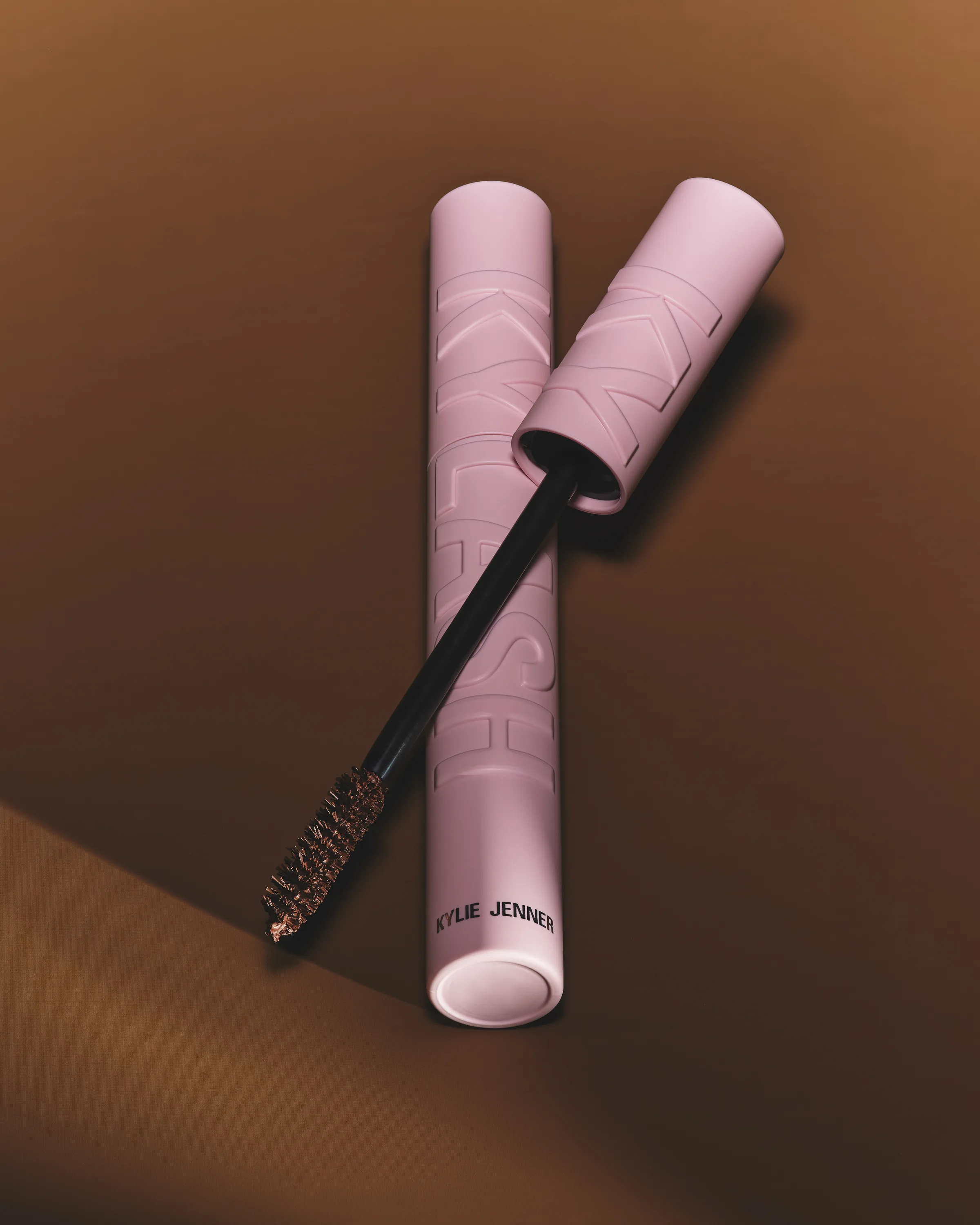 Pink Kylash mascara tube with its brown-tipped wand crossed over it on a warm brown backdrop, for Kylie Cosmetics.