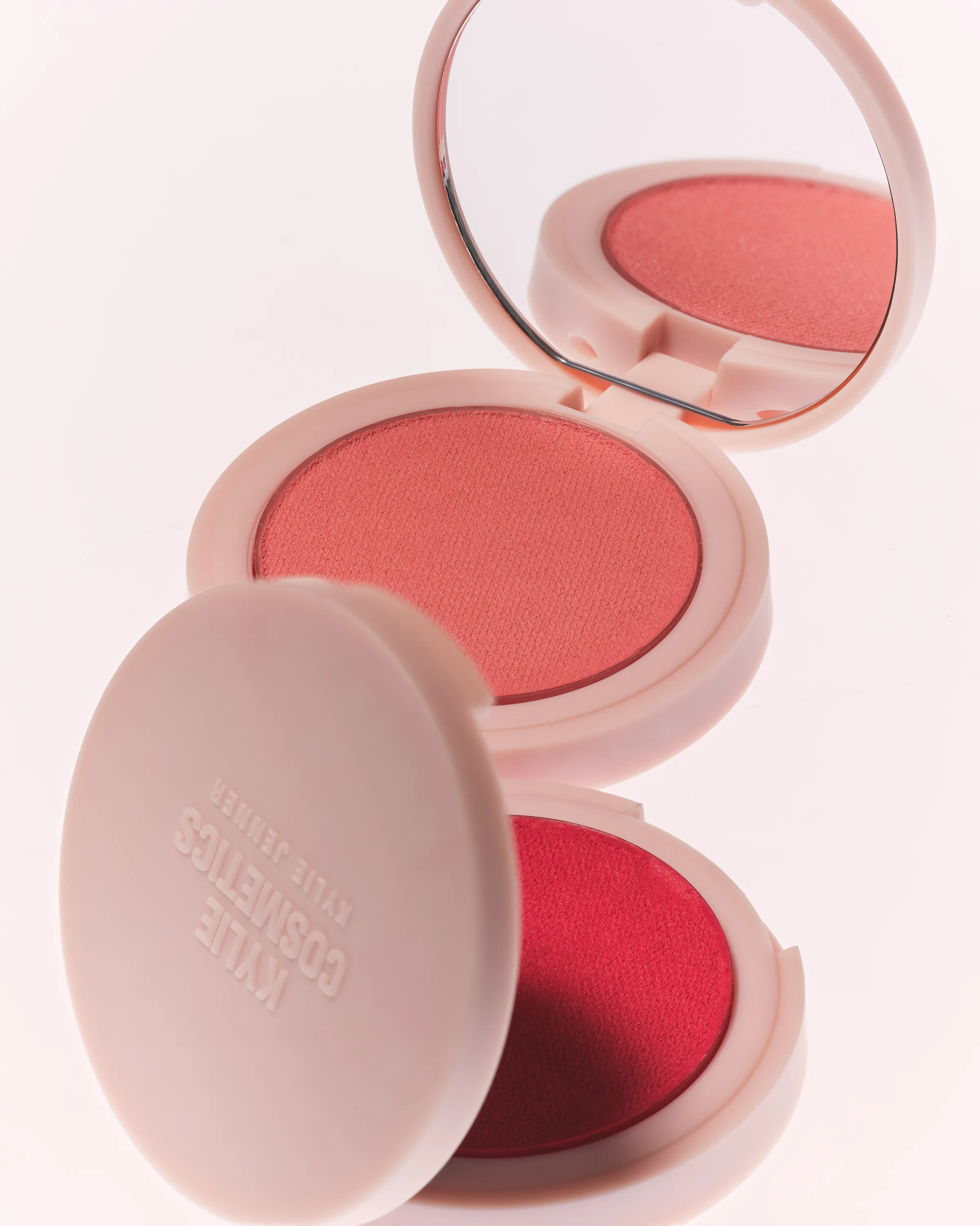 Two open pink blush compacts in coral and red shades with mirror and embossed lid, stacked on a pale backdrop, for Kylie Cosmetics.