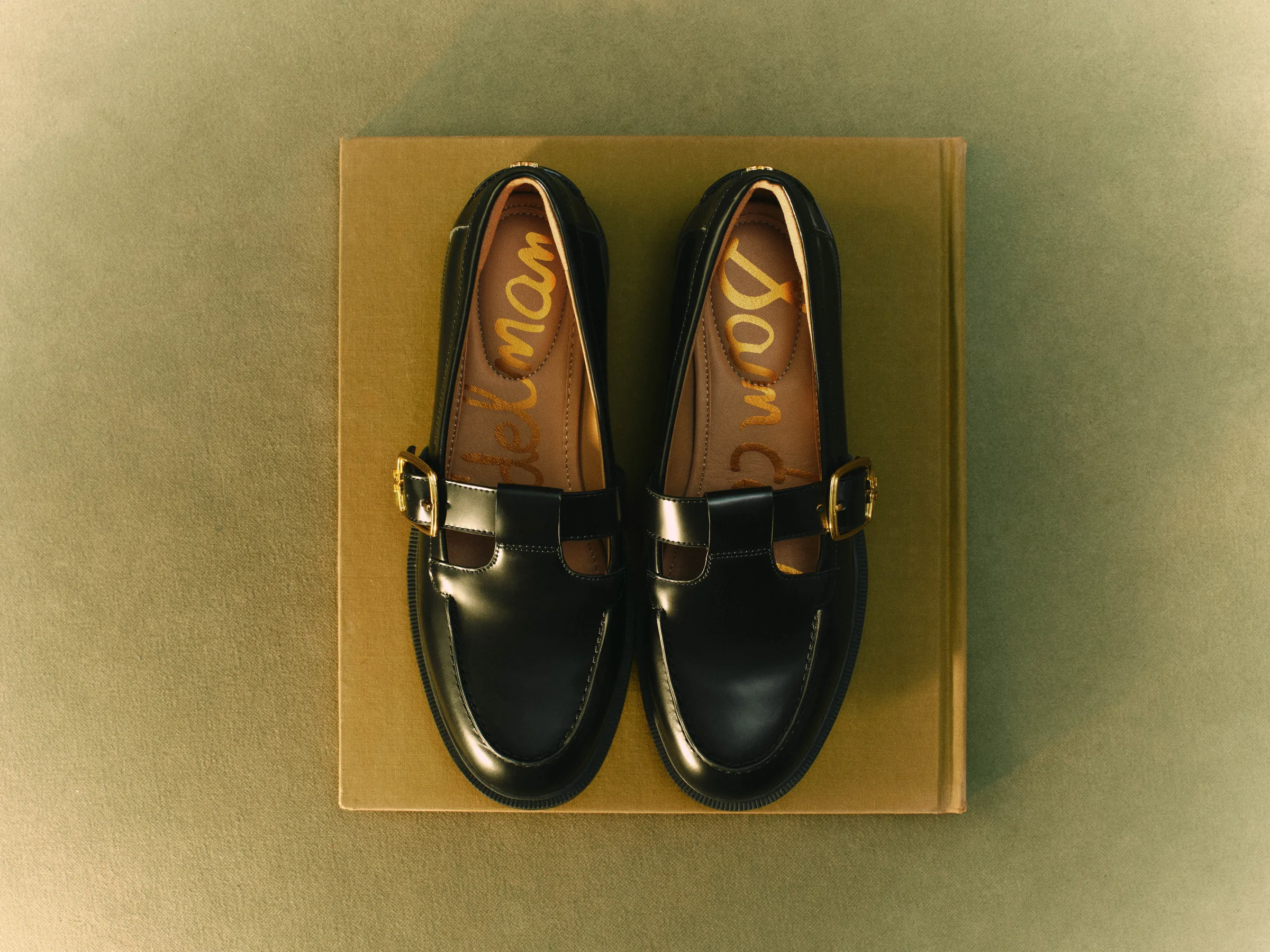 Black patent T-strap loafers with gold buckles set on an ochre cloth book against an olive backdrop, for Sam Edelman.