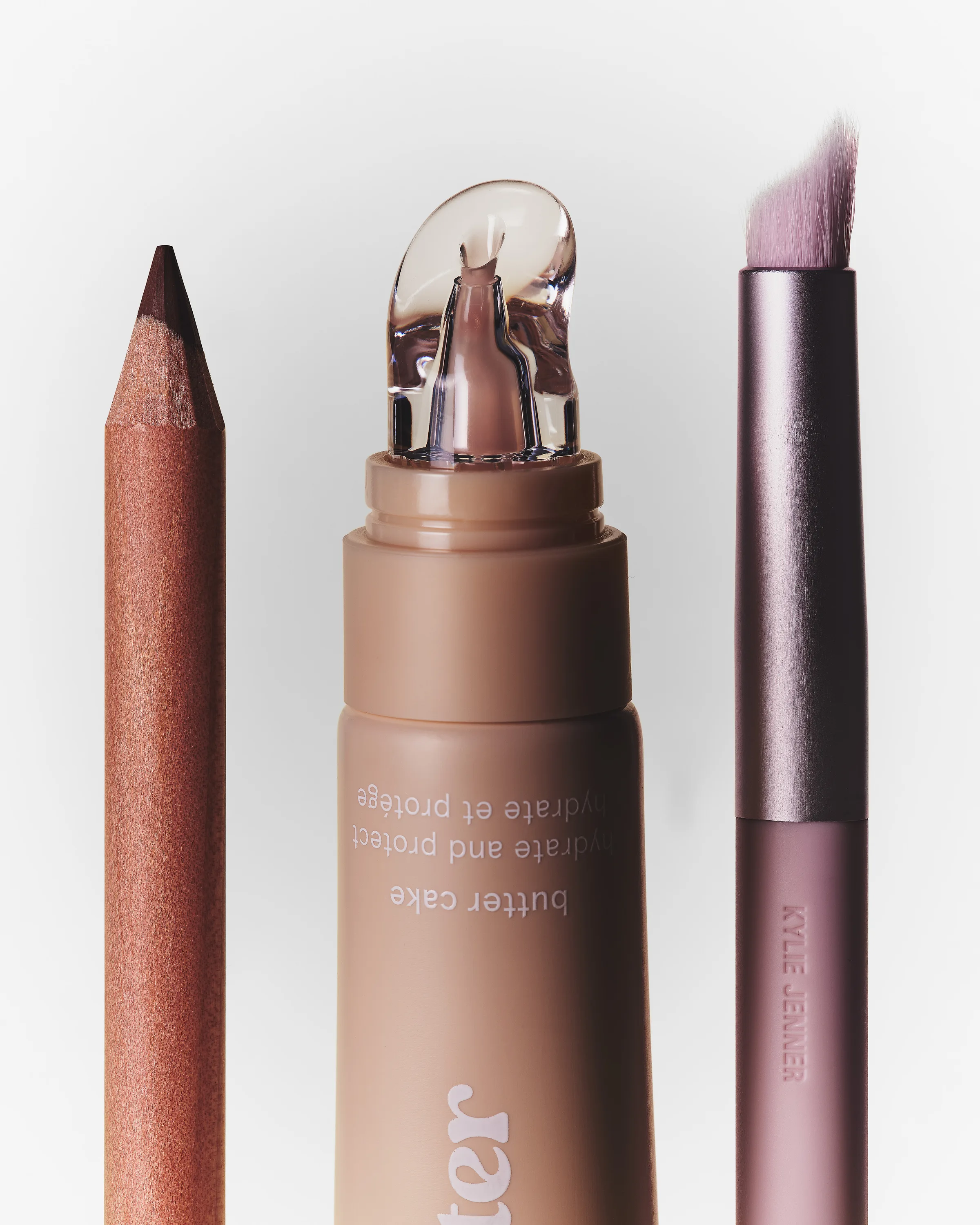 Still life of a nude lip butter tube, copper lip pencil and pink lip brush tips on a pale backdrop, for Kylie Cosmetics.
