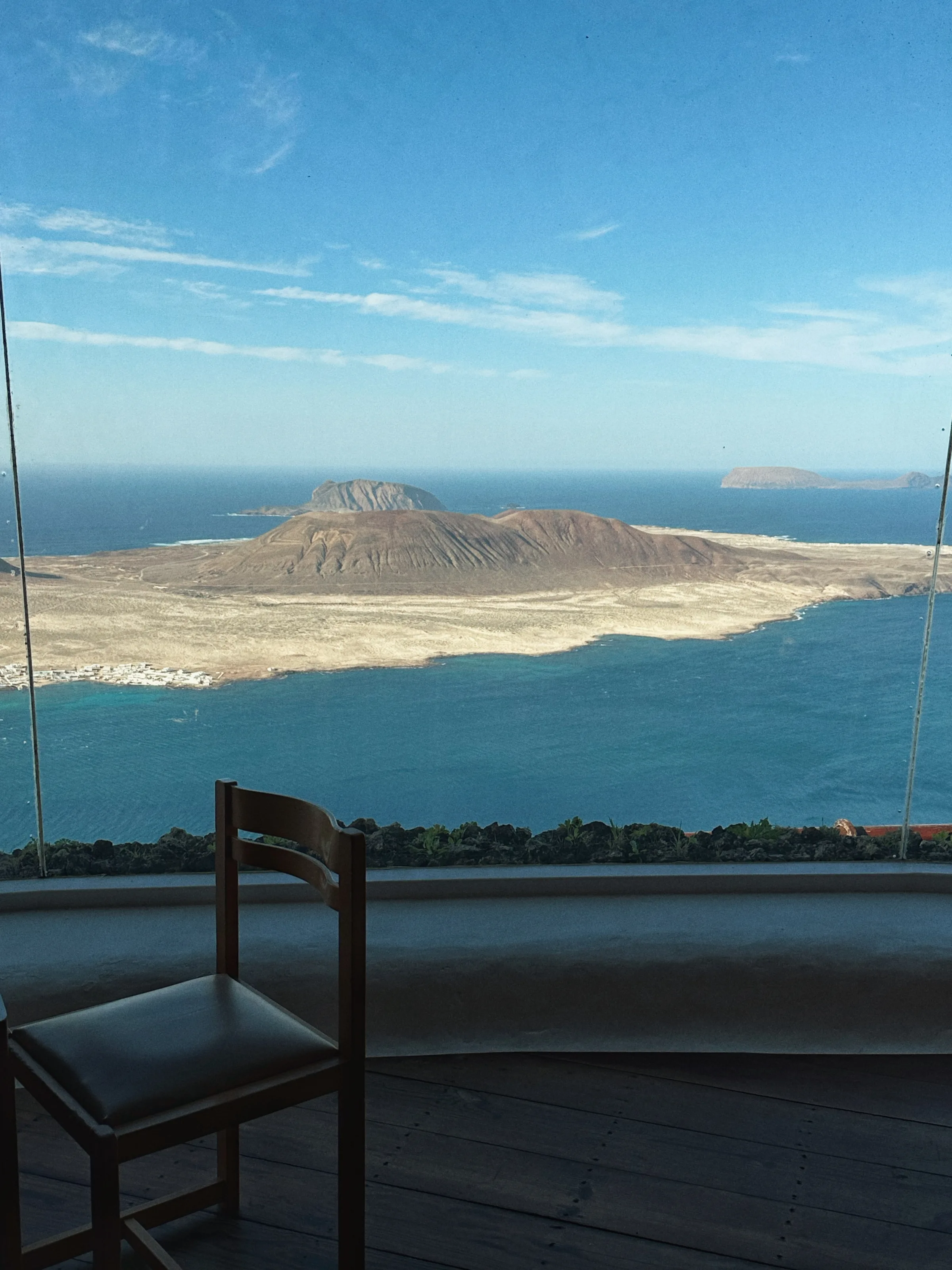 Wooden chair facing a glass window overlooking a volcanic island and blue sea under a clear sky.