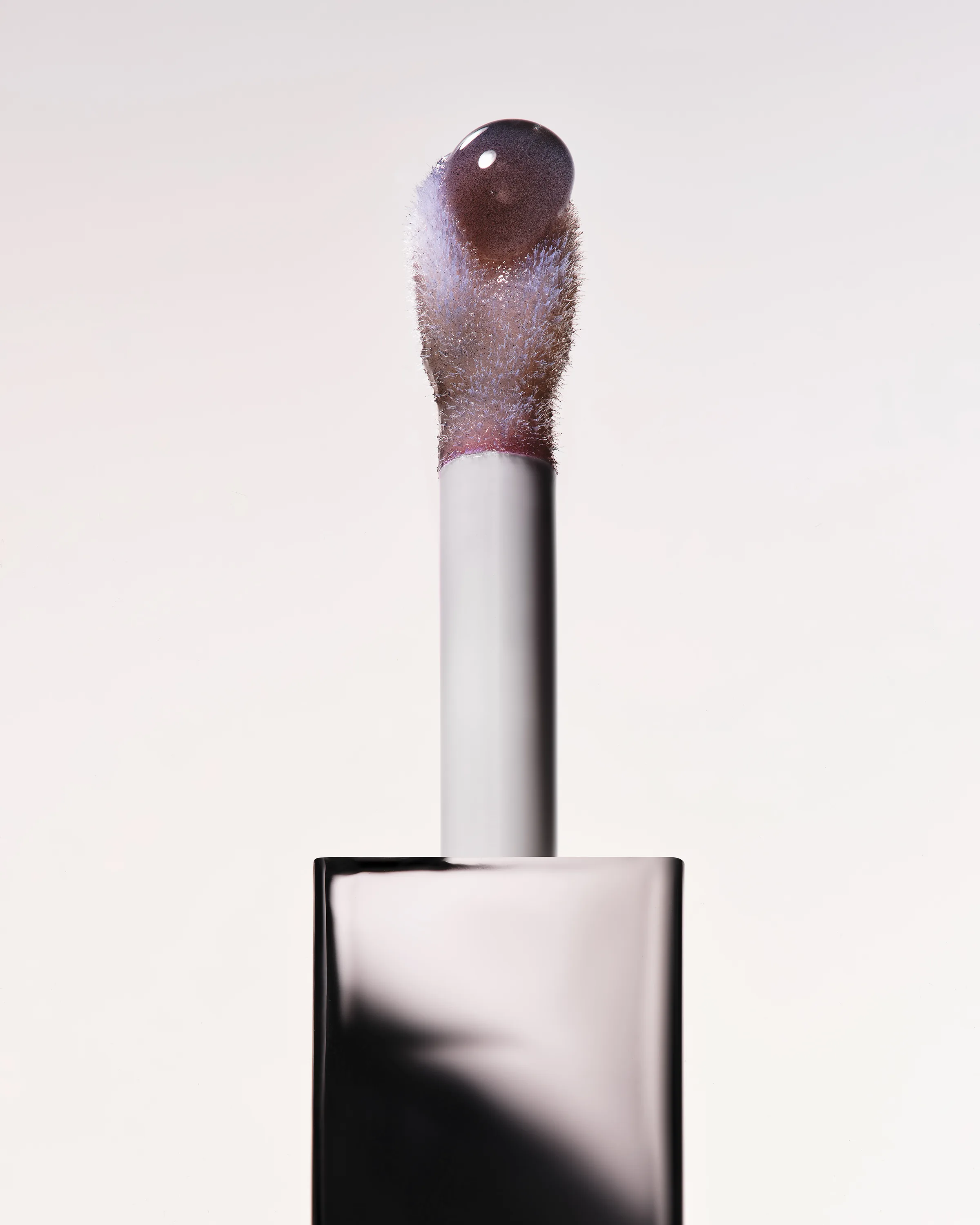 Lip tint doe-foot applicator with a glossy plum drop, rising from a black glossy cap on white, for Kylie Cosmetics.