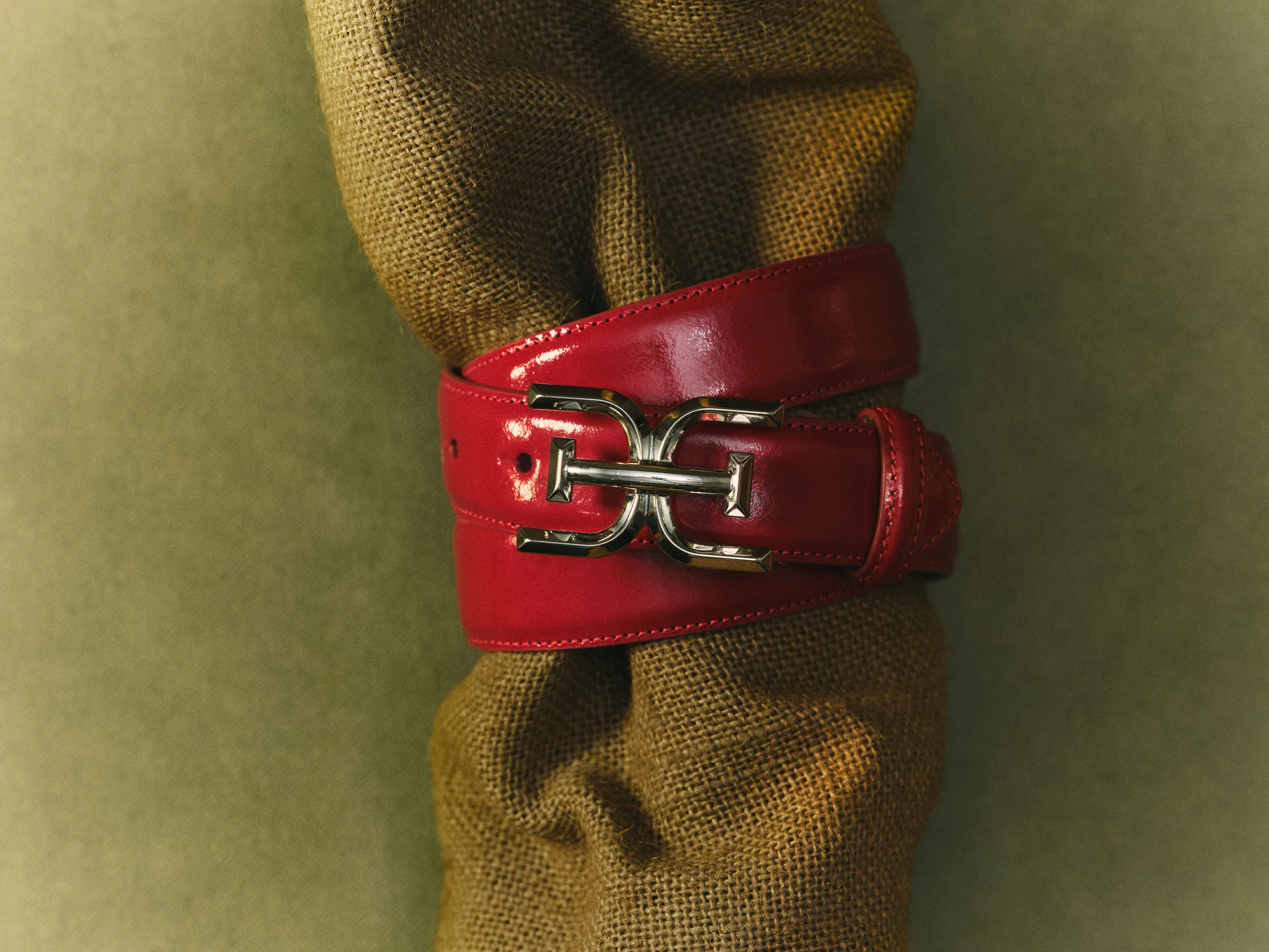 Red patent leather belt with gold double-E buckle wrapped around burlap on an olive backdrop, for Sam Edelman.