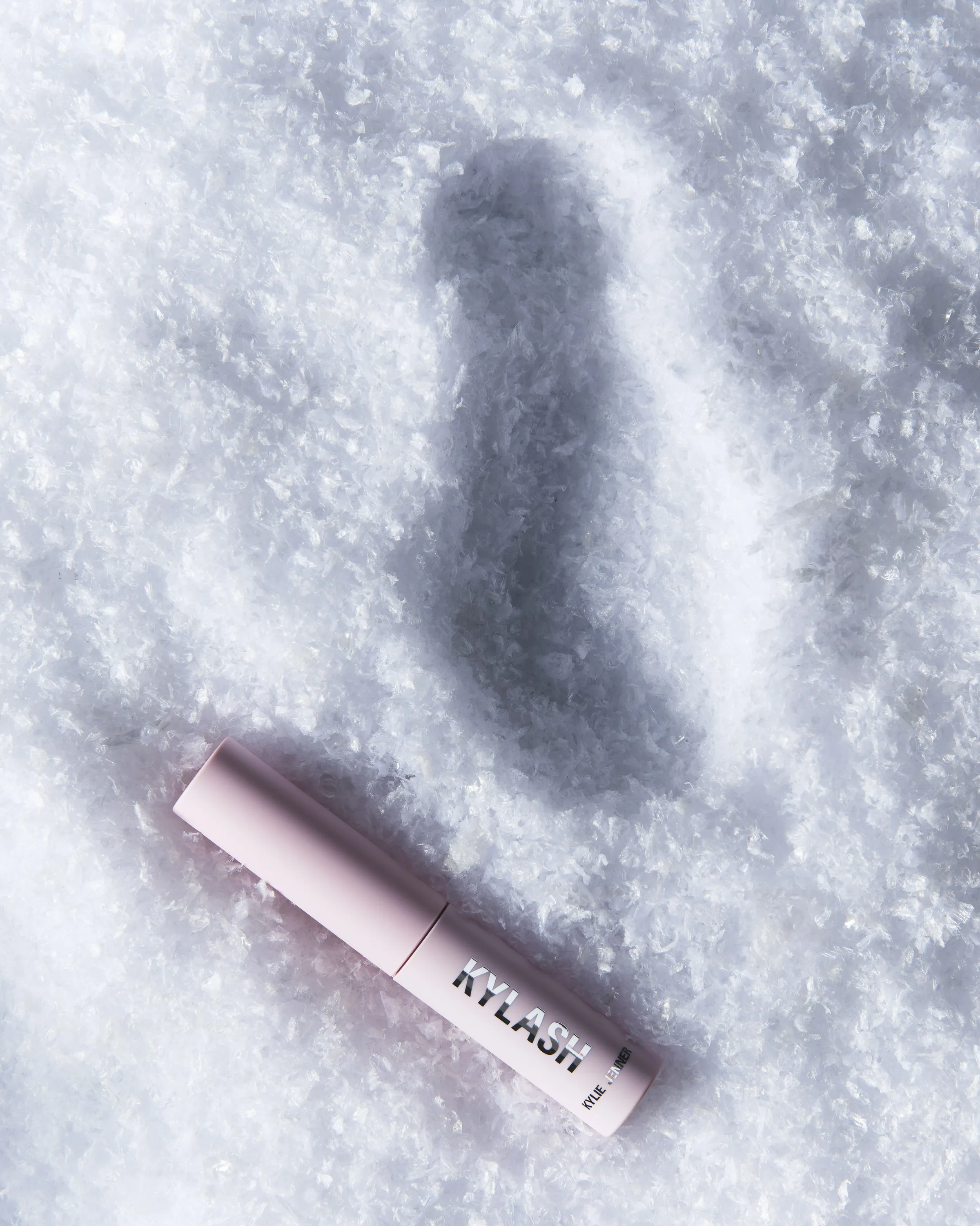 Pink Kylash mini mascara tube lying in snow beside a deep shadow, for Kylie Cosmetics.