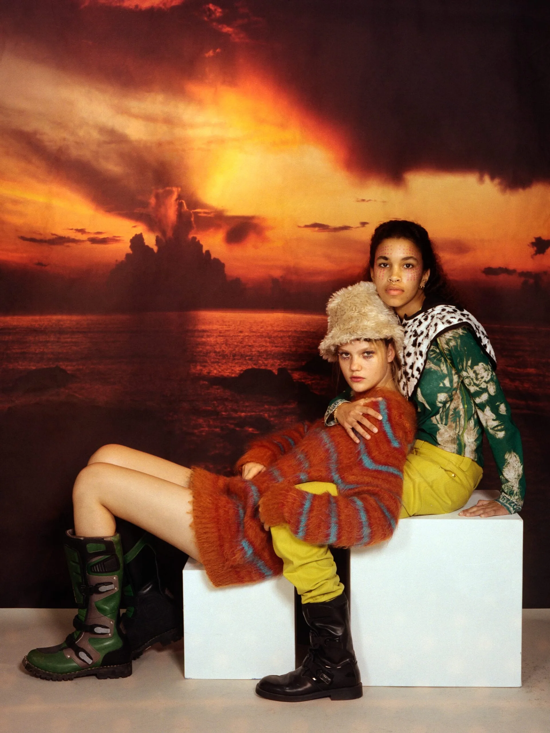 Two models in a fuzzy striped sweater, fur hat and green floral top pose on white cubes before a sunset backdrop.