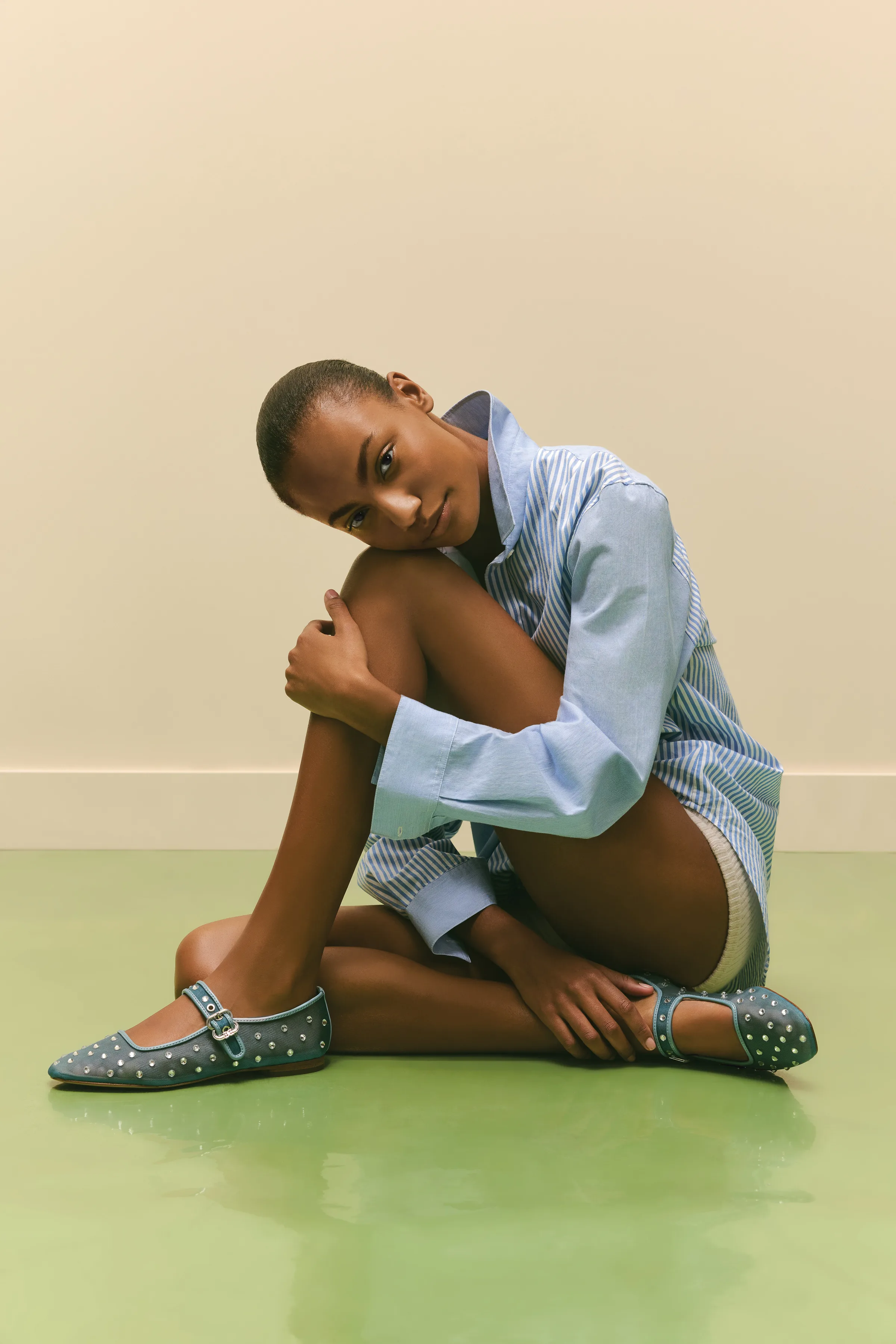 Model in a striped blue shirt sits on a green floor wearing studded denim Mary Jane flats, for Sam Edelman.