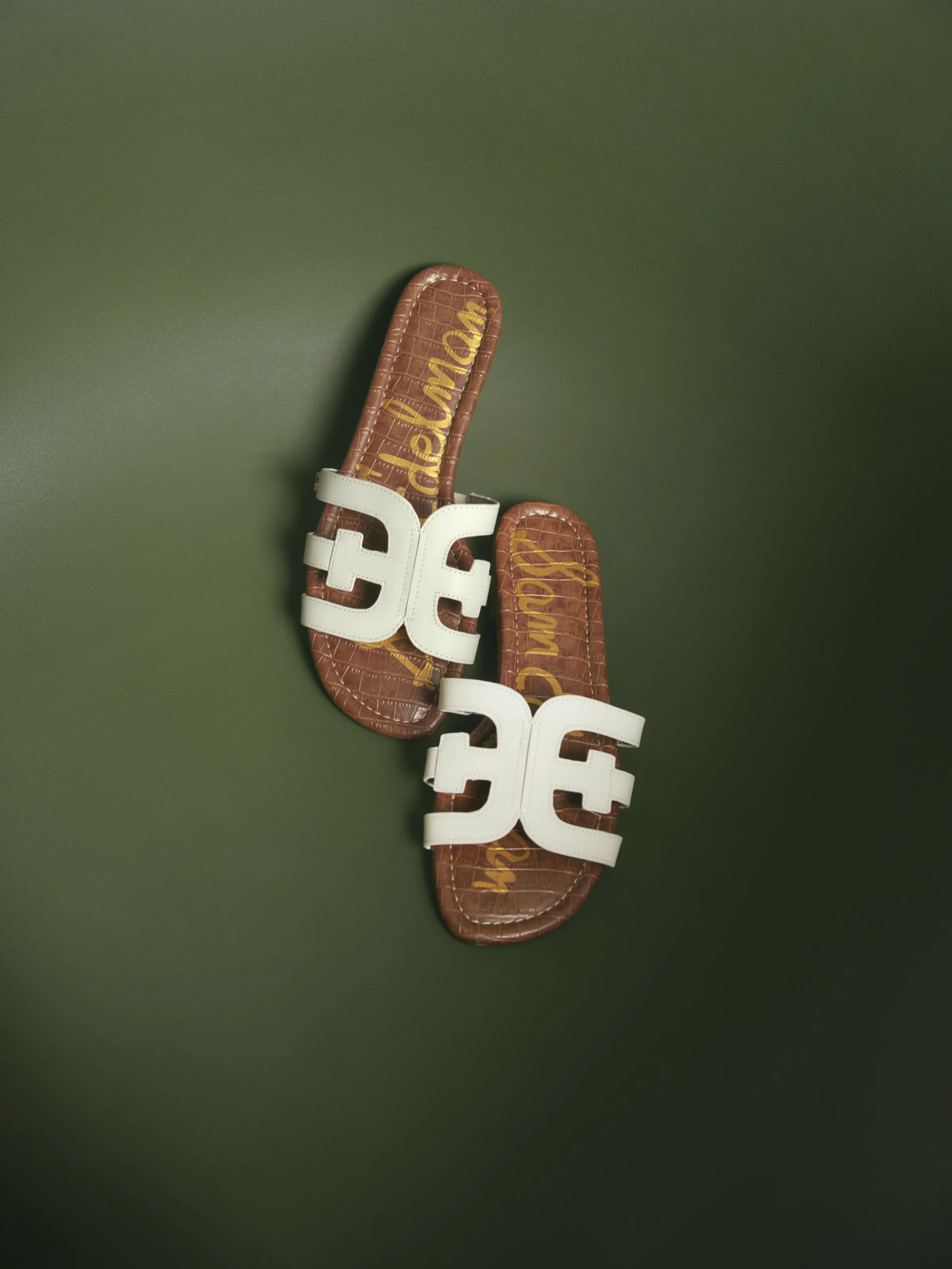Pair of white double-E slide sandals with tan croc-embossed footbeds on an olive green backdrop, for Sam Edelman.