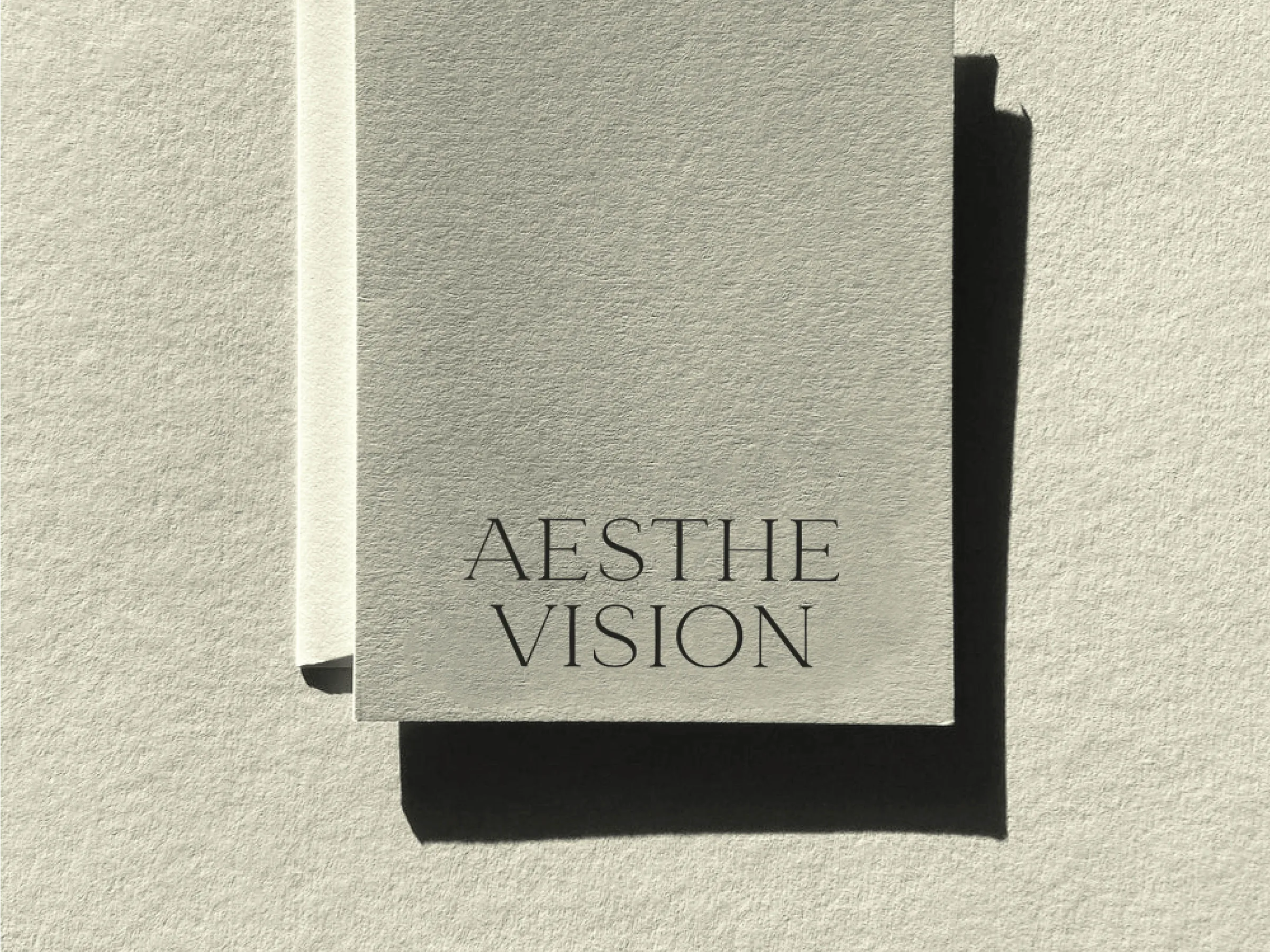 Pale grey textured card printed with the Aesthe Vision wordmark, casting a hard shadow on paper, for Aesthevision.