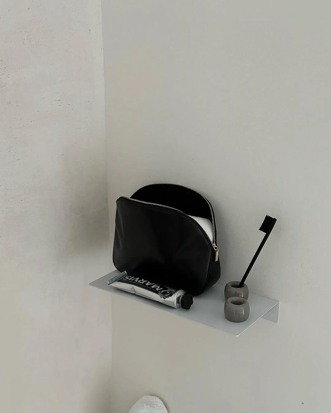 Black zip toiletry bag on a white wall shelf beside Marvis toothpaste and a black toothbrush in a stone holder.