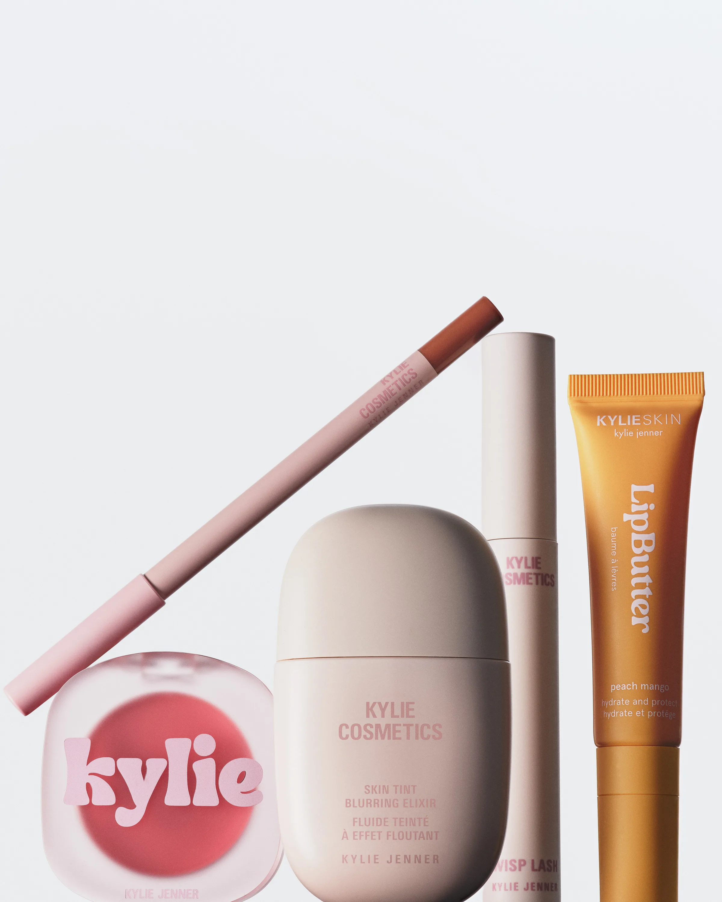 Group of pink blush, lip liner, skin tint, mascara and peach mango Lip Butter on a white backdrop, for Kylie Cosmetics.