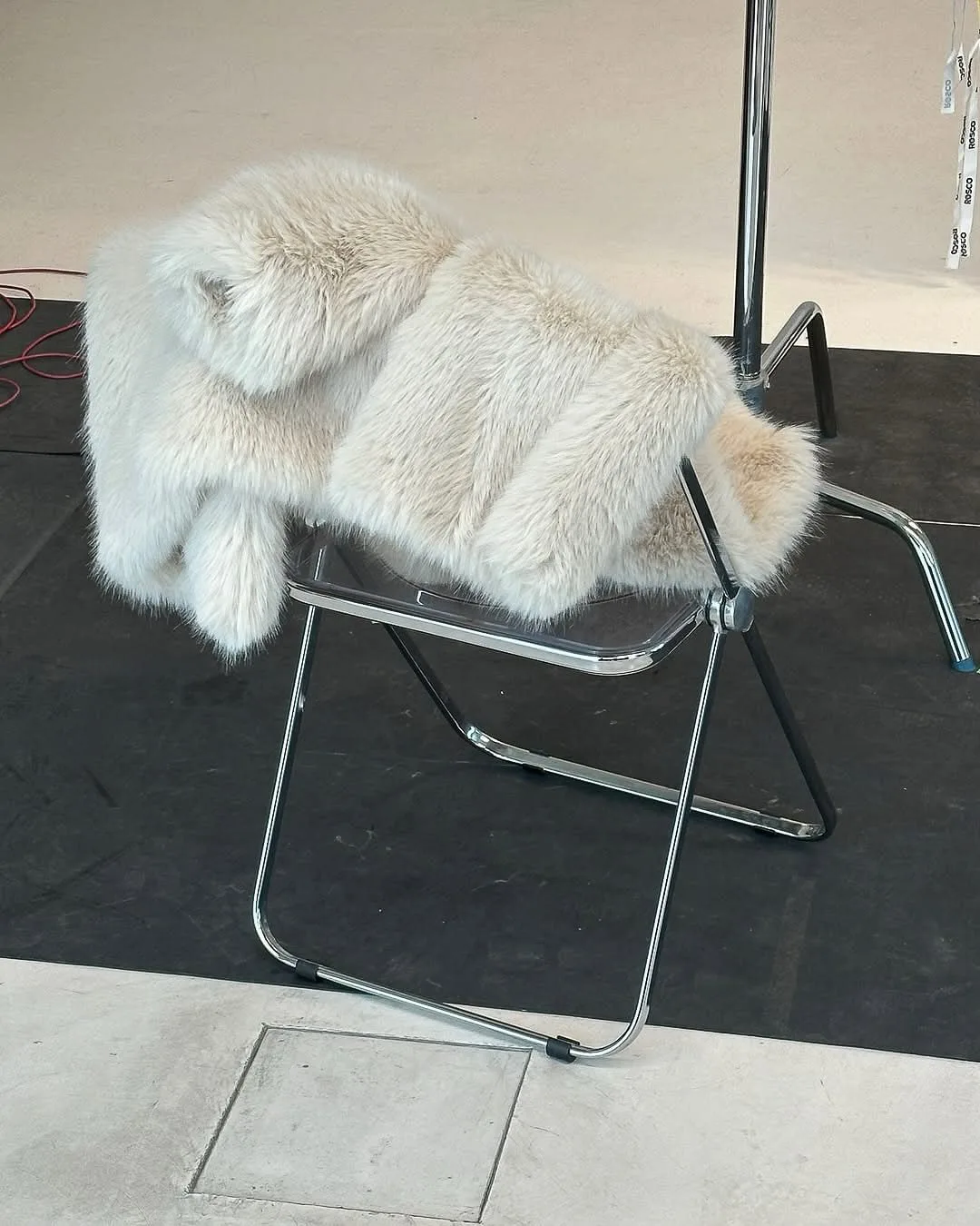 Cream faux fur coat draped over a clear acrylic folding chair on a studio floor, for Aina Velasco.