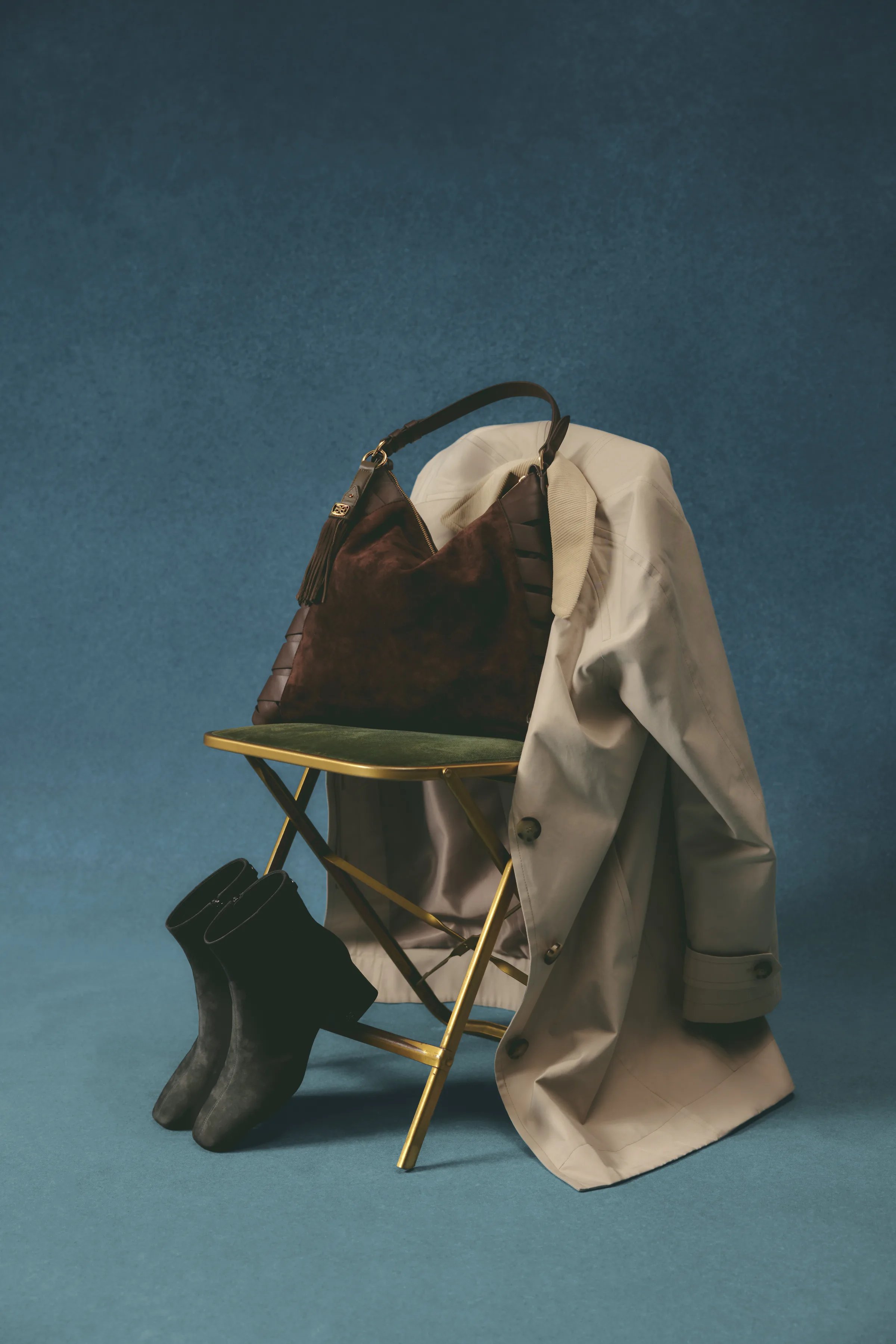 Grey suede ankle boots beside a brass folding chair draped with a beige trench coat and brown suede bag, for Sam Edelman.