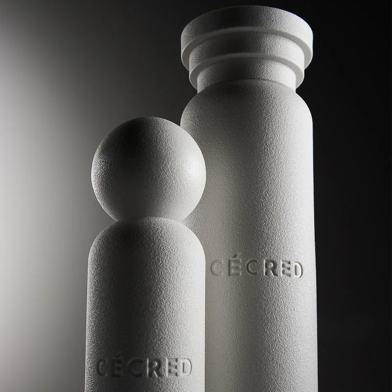 Two textured white hair care bottles with embossed logos, one with a sphere cap, lit dramatically on grey, for Cécred.