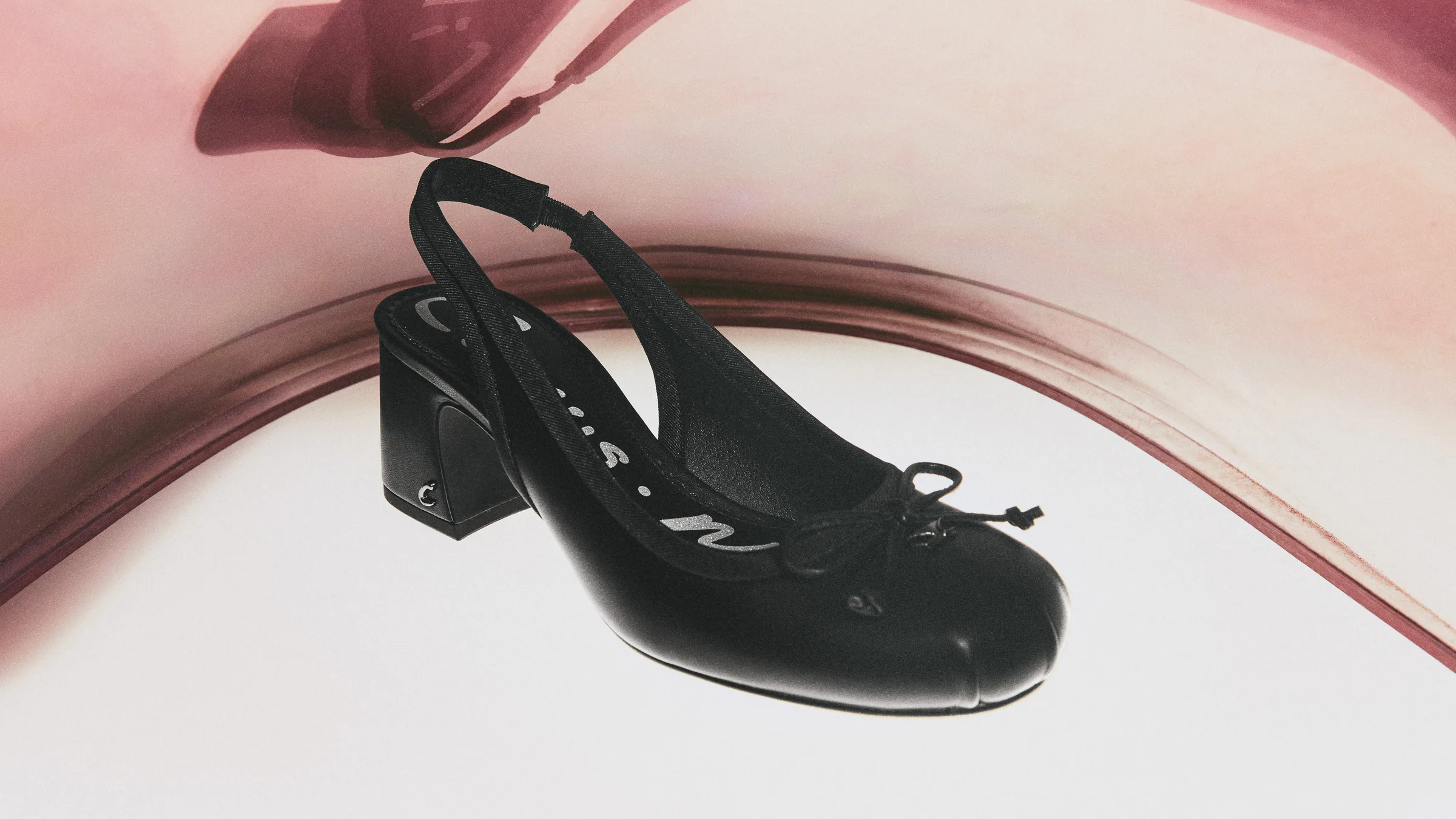 Black leather slingback pump with bow and block heel floating over a curved pink and white backdrop, for Circus NY.