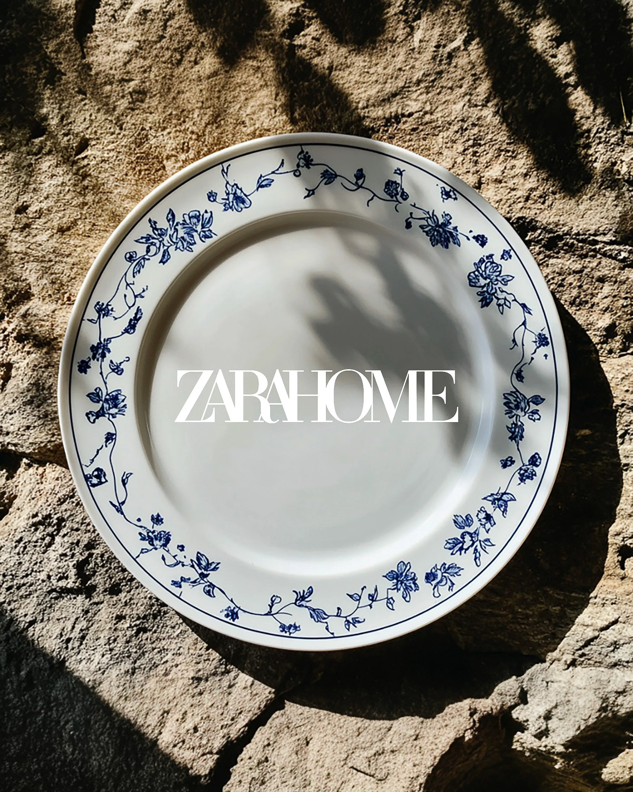 AI image of a white plate with blue floral border on sunlit stone with leaf shadows, Zara Home logo overlaid. Zara Home AI Concept.